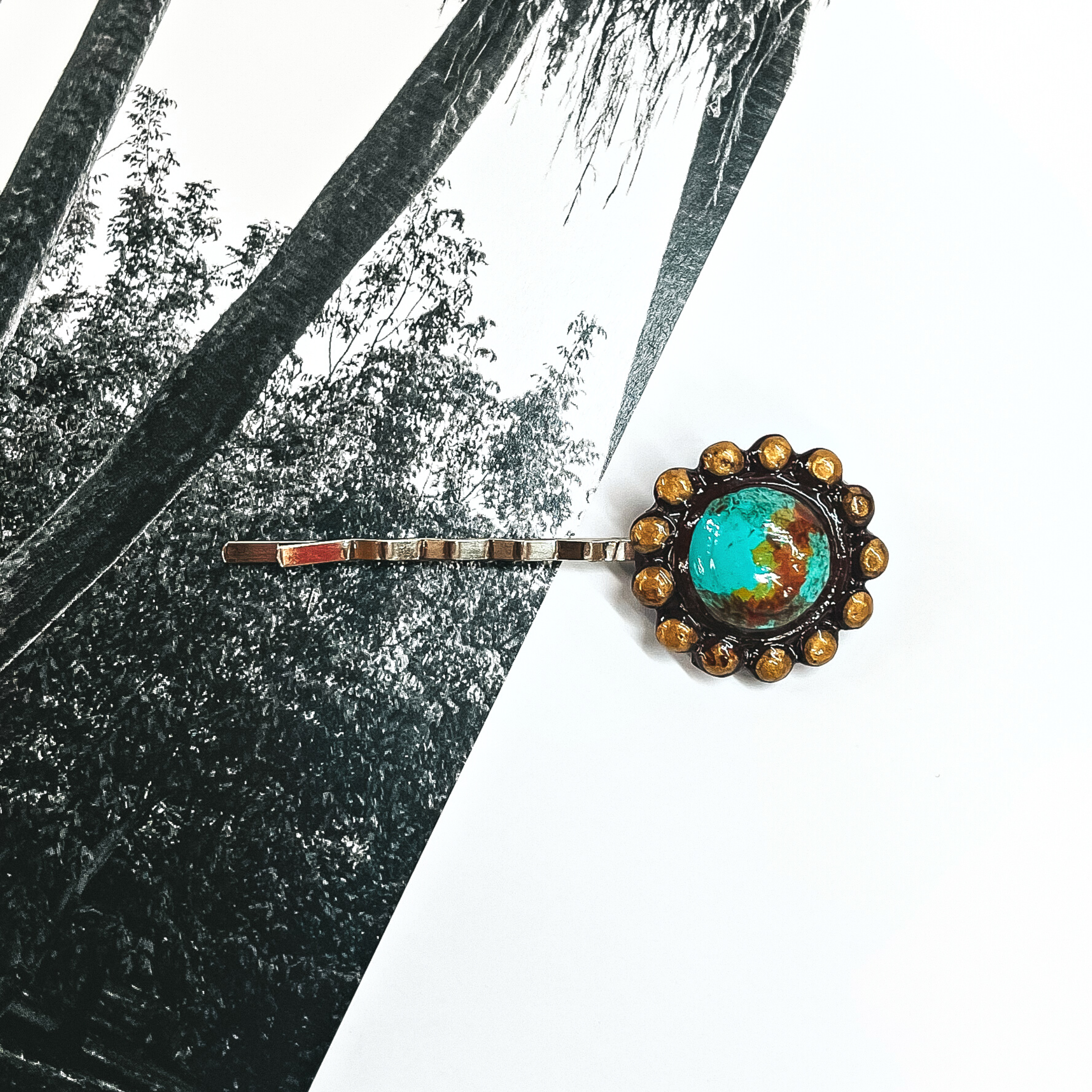 This is a silver tone bobby pin with a clay piece at the end of the pin. It is a circle with rounded ridges, the circle is teal/turquoise and brown. The rounded ridges are painted gold. This bobby pin is laying on a black and white outdoor book page,