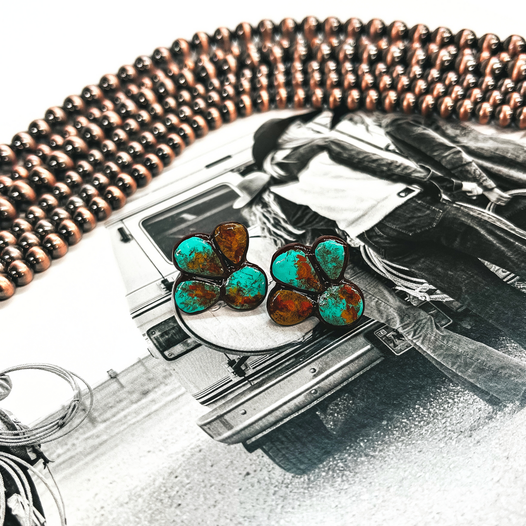 These are a pair of clay earrings in teal/turquoise and brown. They havve a circle with three petals in the side. These earrings are placed on an outdoor black and white book page with copper beads in the back as decor. 