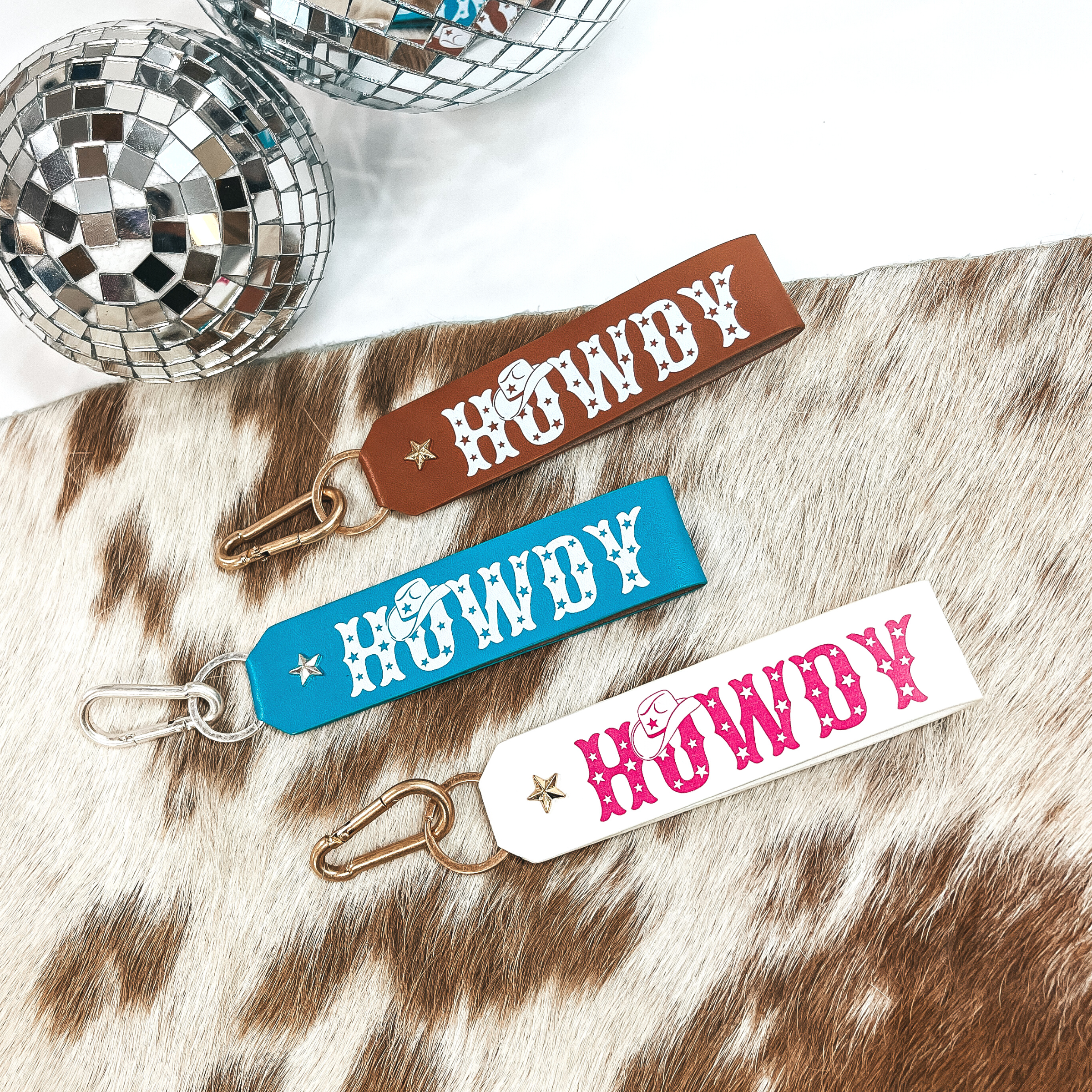 There are three leather keychains in brown/tan, turquoise, and white with  hot pink. They all say, Howdy, with uppercase letters and star print on the  letters, and a wobody hat on top of the, O. All keychain's are laying on a  white and brown cow hide and on a white background with two silver disco balls  in the back as decor.