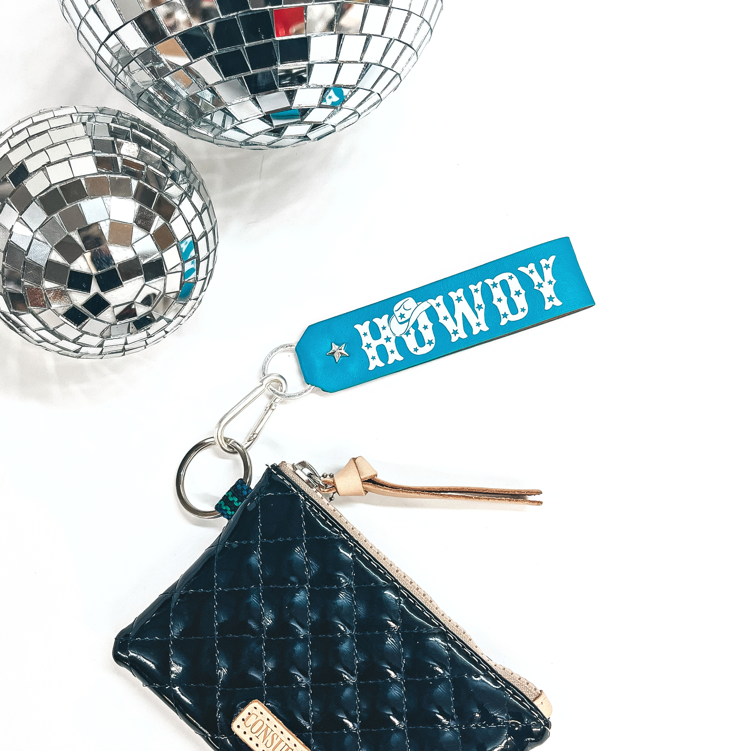 This is a turquoise colored leather keychain with a matte silver key ring.  The keychain has says, Howdy, in uppercase with star print inside and a  cowboy hat on top of the, O.  It's connected to a black shiny Consuela pouch and on a white background with  two disco balls in the back as decor.