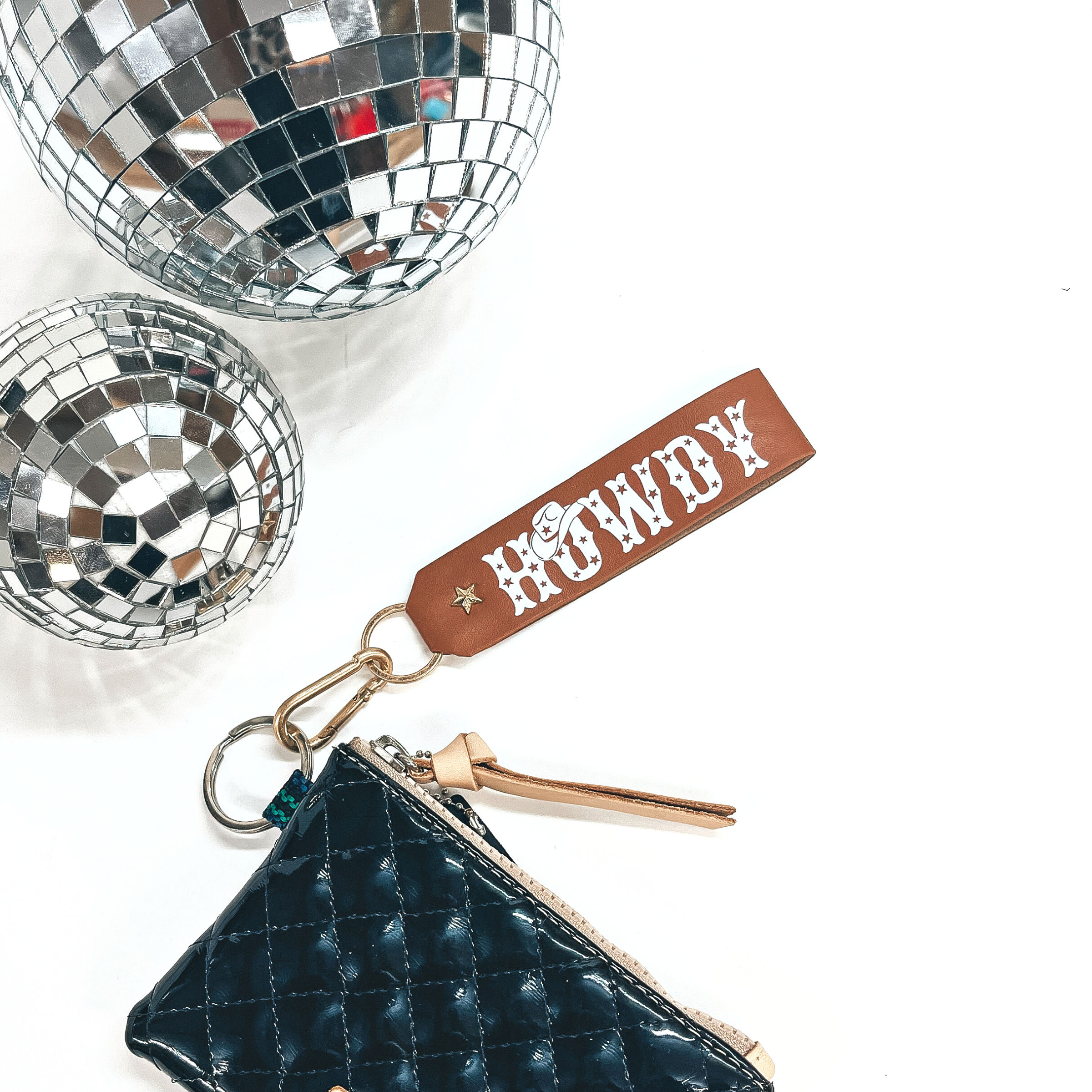 This is a brown/tan colored leather keychain with a matte gold key ring.  The keychain says, Howdy, in uppercase with star print inside and a  cowboy hat on top of the, O, in white. It's connected to a black shiny Consuela pouch and on a white background with  two disco balls in the back as decor.