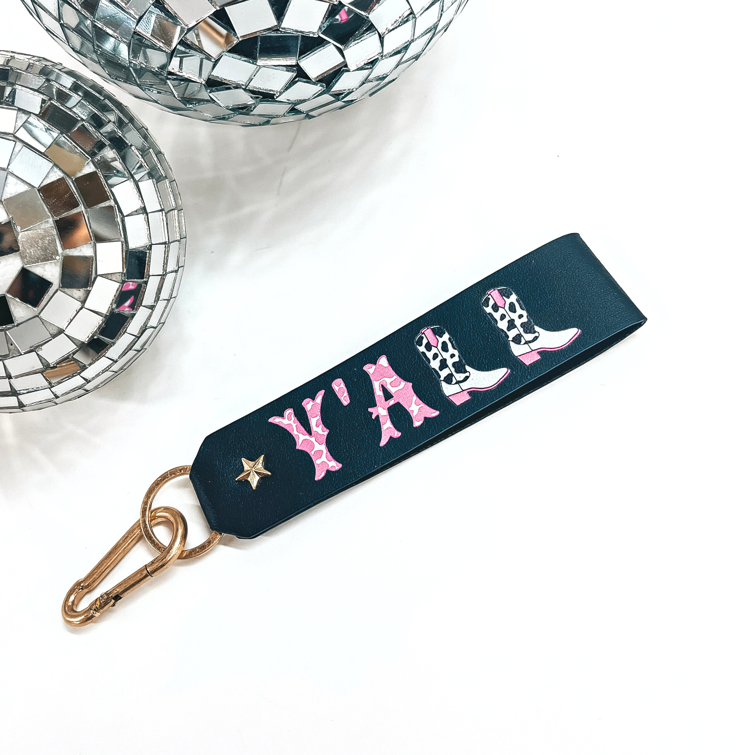 This is a black leather keychain with a matte gold key ring and a star.  It says, 'Y'ALL', with pink and white cow print inside the letters, there is  also two boots as L's. The boots have black and white cow print with pink  detailing. This keychain is taken on a white background with two disco balls  in the back as decor.