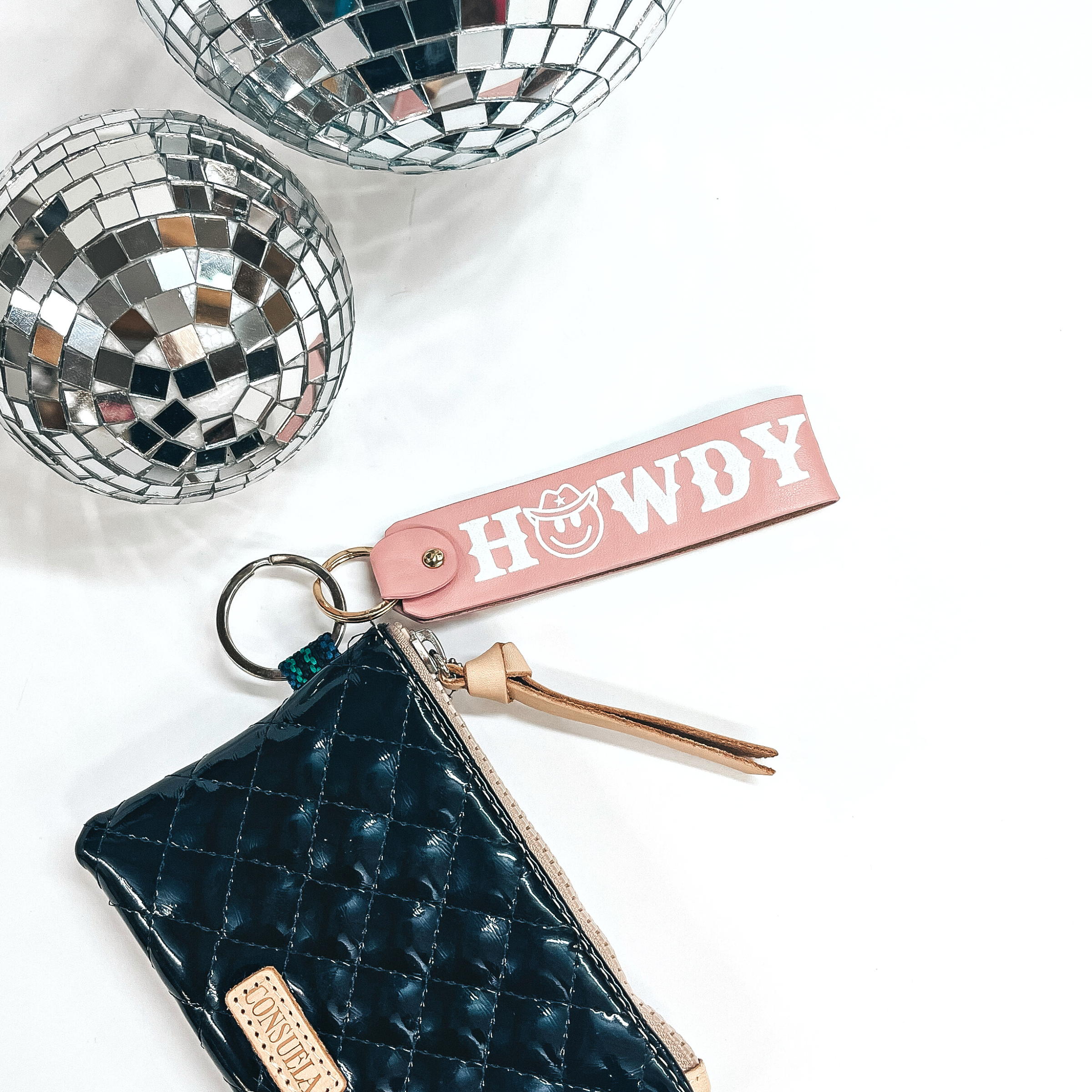 This is a light pink leather keychain with a matte gold key ring. It says, 'Howdy', with uppercase white lettering and it has a happy face with a  cowboy hat as the, O. This keychain is connected to a black shiny Consuela  pouch and on a white background, with two disco balls in the back as decor.