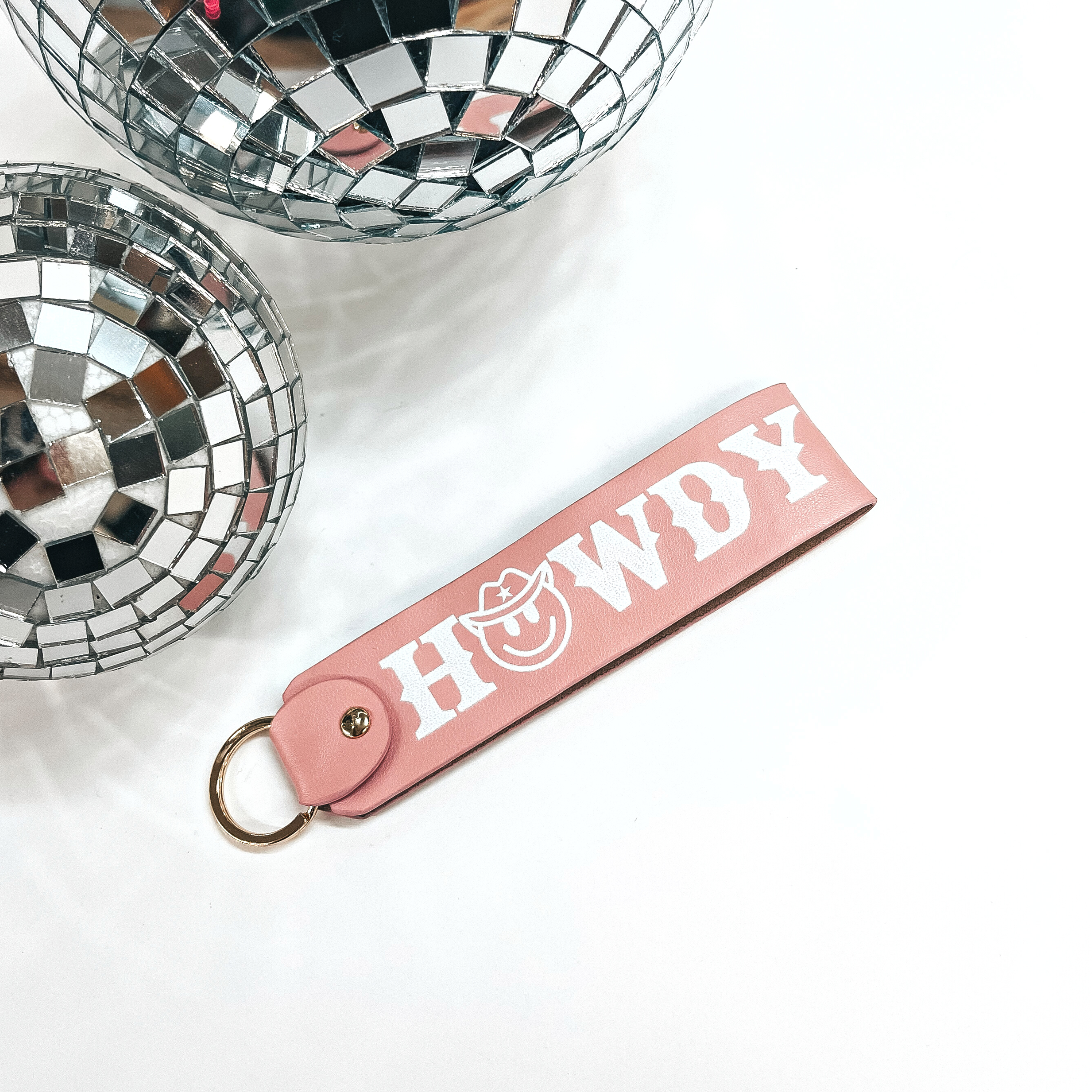 This is a light pink leather keychain with a matte gold key ring. It says, 'Howdy', with uppercase white lettering and it has a happy face with a  cowboy hat as the, O. This keychain is taken on a white background with two  disco balls in the back as decor.