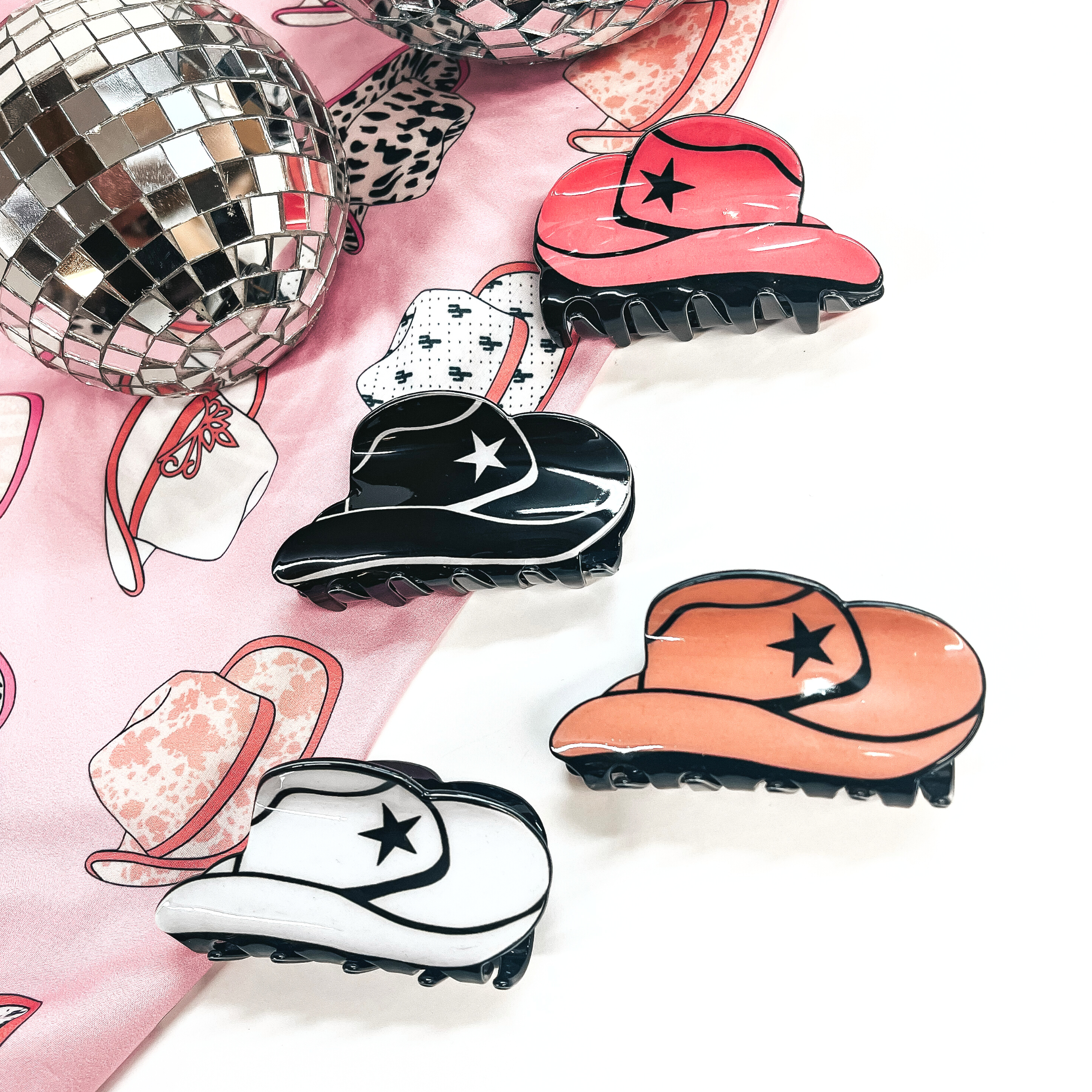 There are four solid color cowboy hat hair clips laying on a cowboy hat print fabric and a white background with two disco balls in the back as decor.