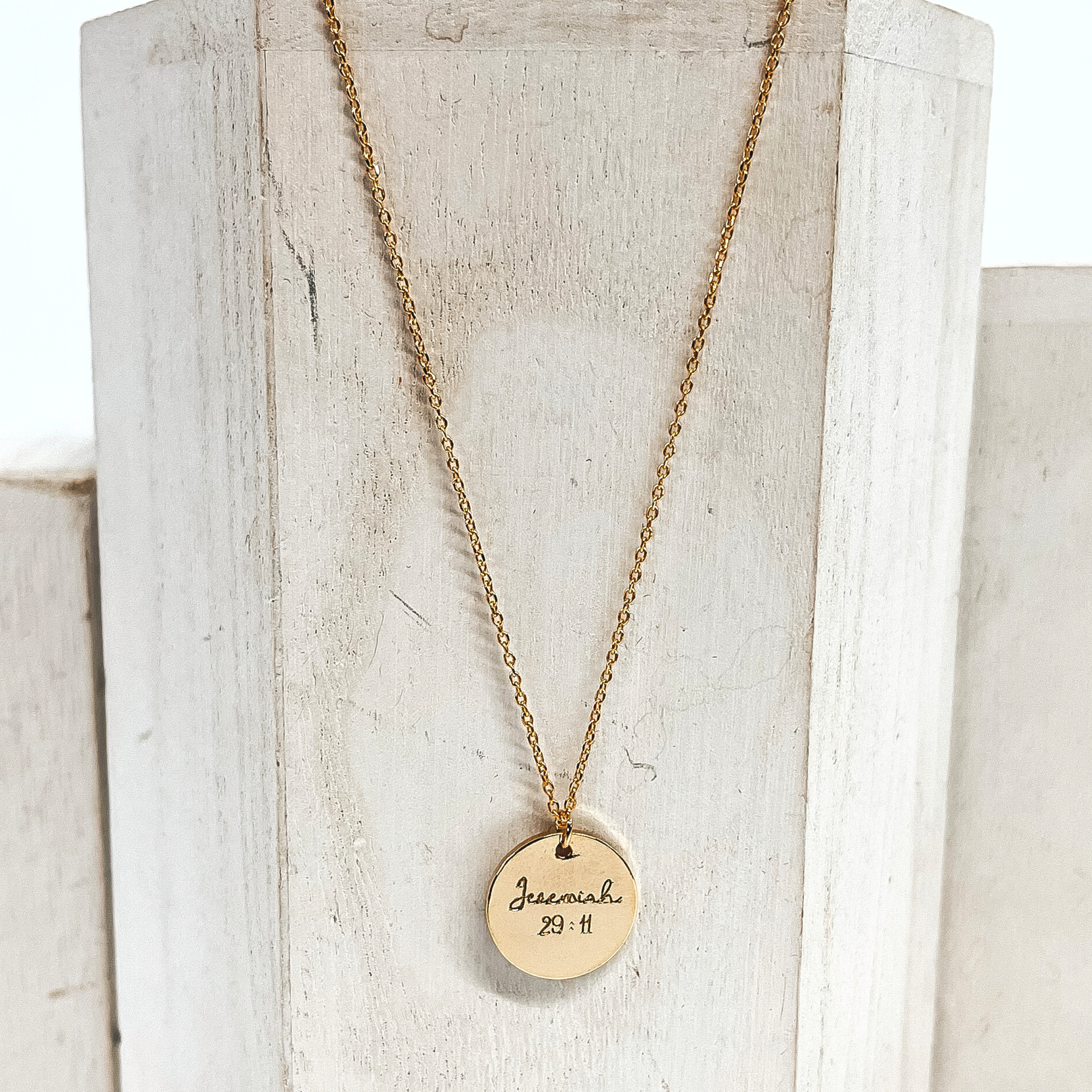 This is a gold thin necklace with a circle pendant that says, Jeremiah 29:11. This necklace is placed on a white block.