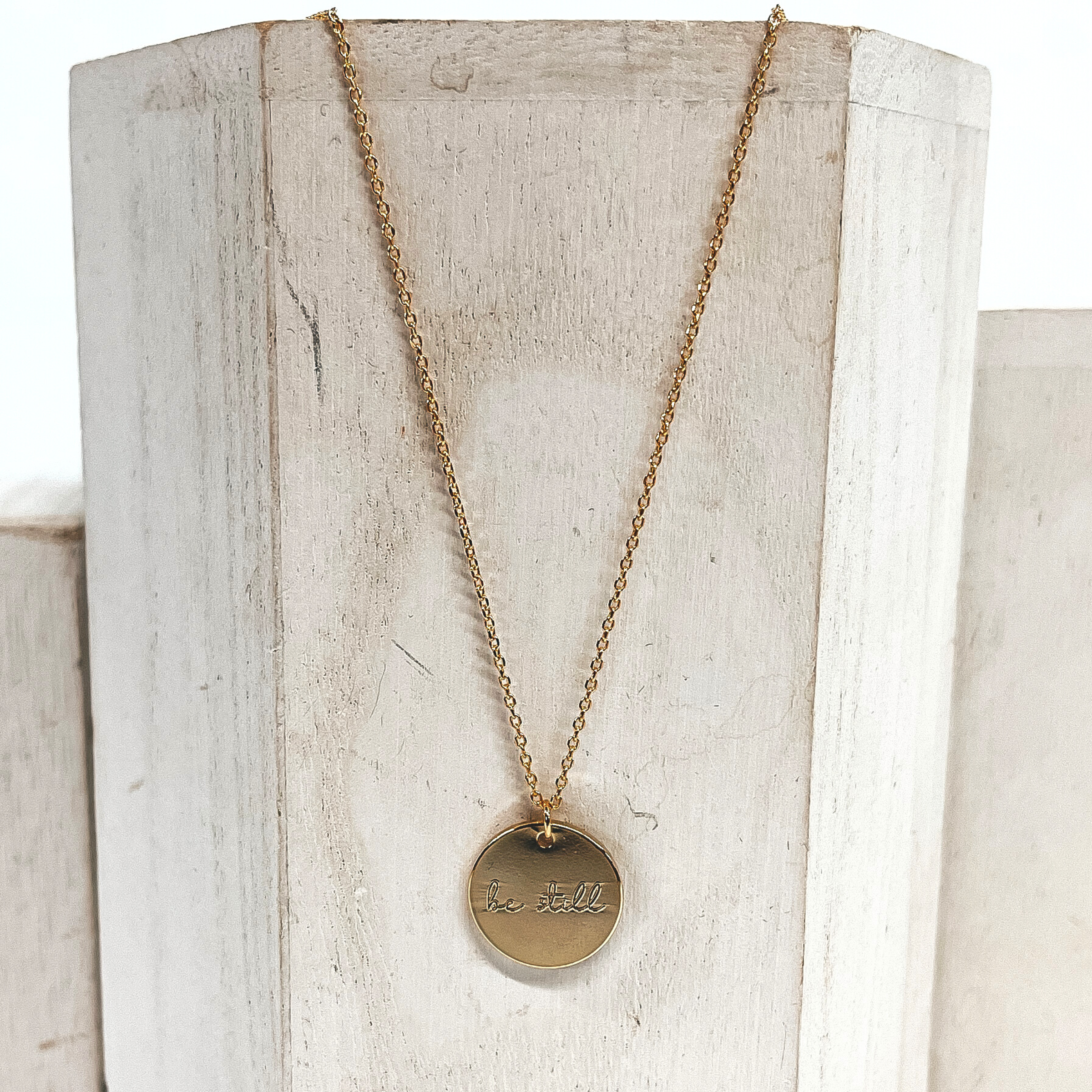 This is a thin gold necklace with a circle pendant that says, Be Still. This necklace is placed on a white/ivory block.