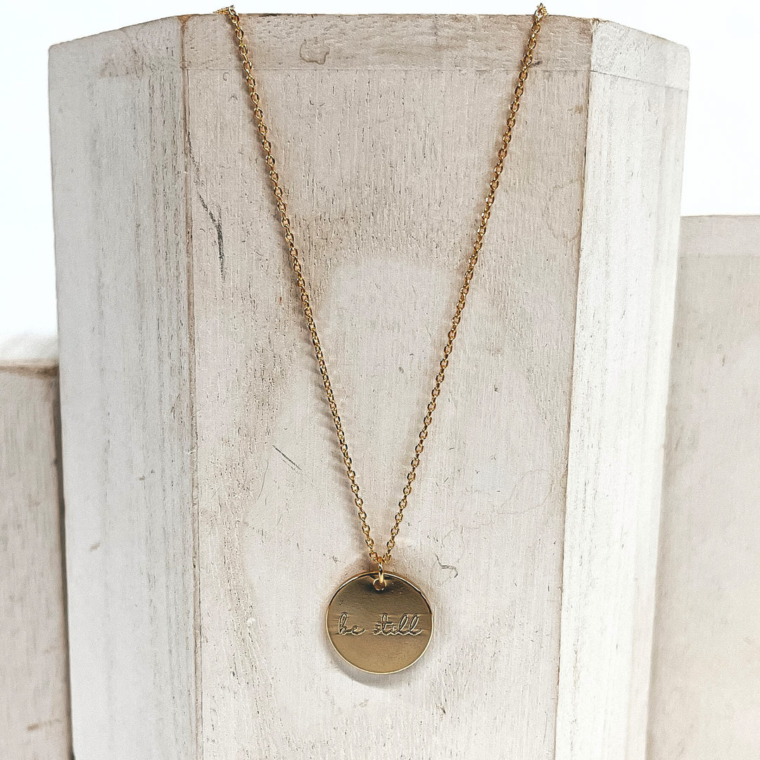 This is a thin gold necklace with a circle pendant that says, Be Still. This necklace is placed on a white/ivory block.