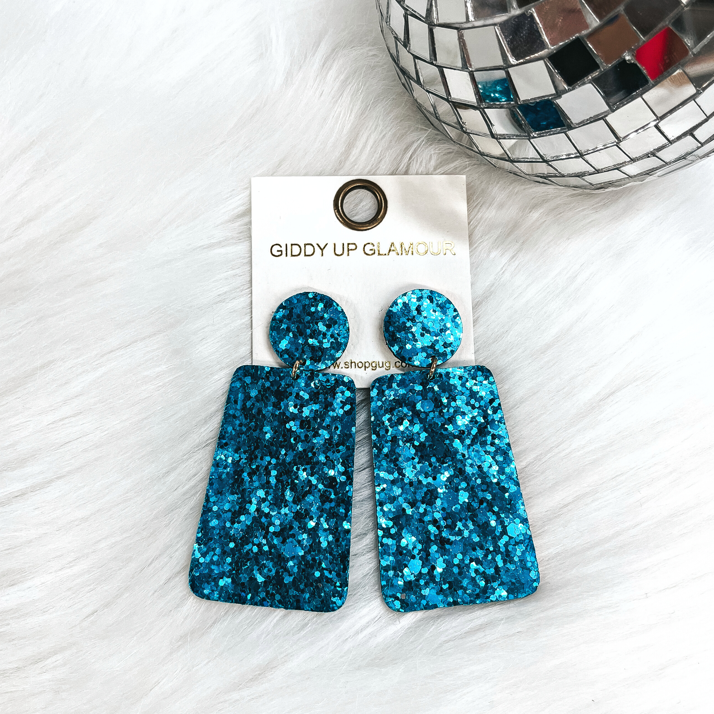These are rectangle glitter drop earrings in turquoise, the postback is a circle  with a small gold tone connector and a rectangle drop. These earrings  are laying on white fur with a small disco ball in the back as decor.
