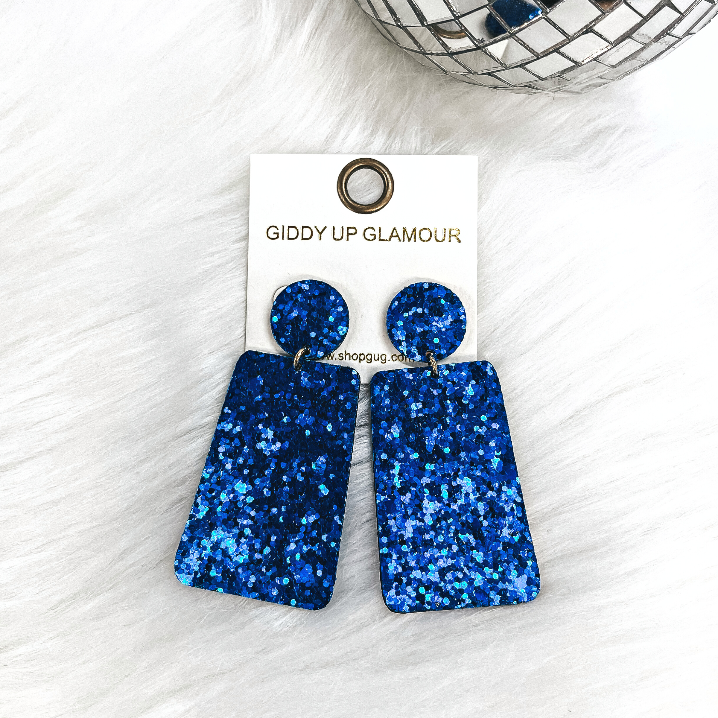 These are rectangle glitter drop earrings in blue, the postback is a circle  with a small gold tone connector and a rectangle drop. These earrings  are laying on white fur with a small disco ball in the back as decor.