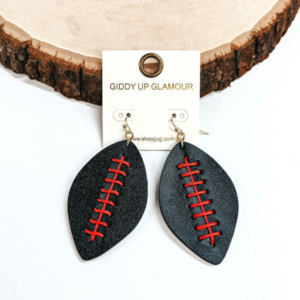 These are football shaped earrings in black with red stitching in the center. These earrings are leaning up against a slab of wood and on a white background.