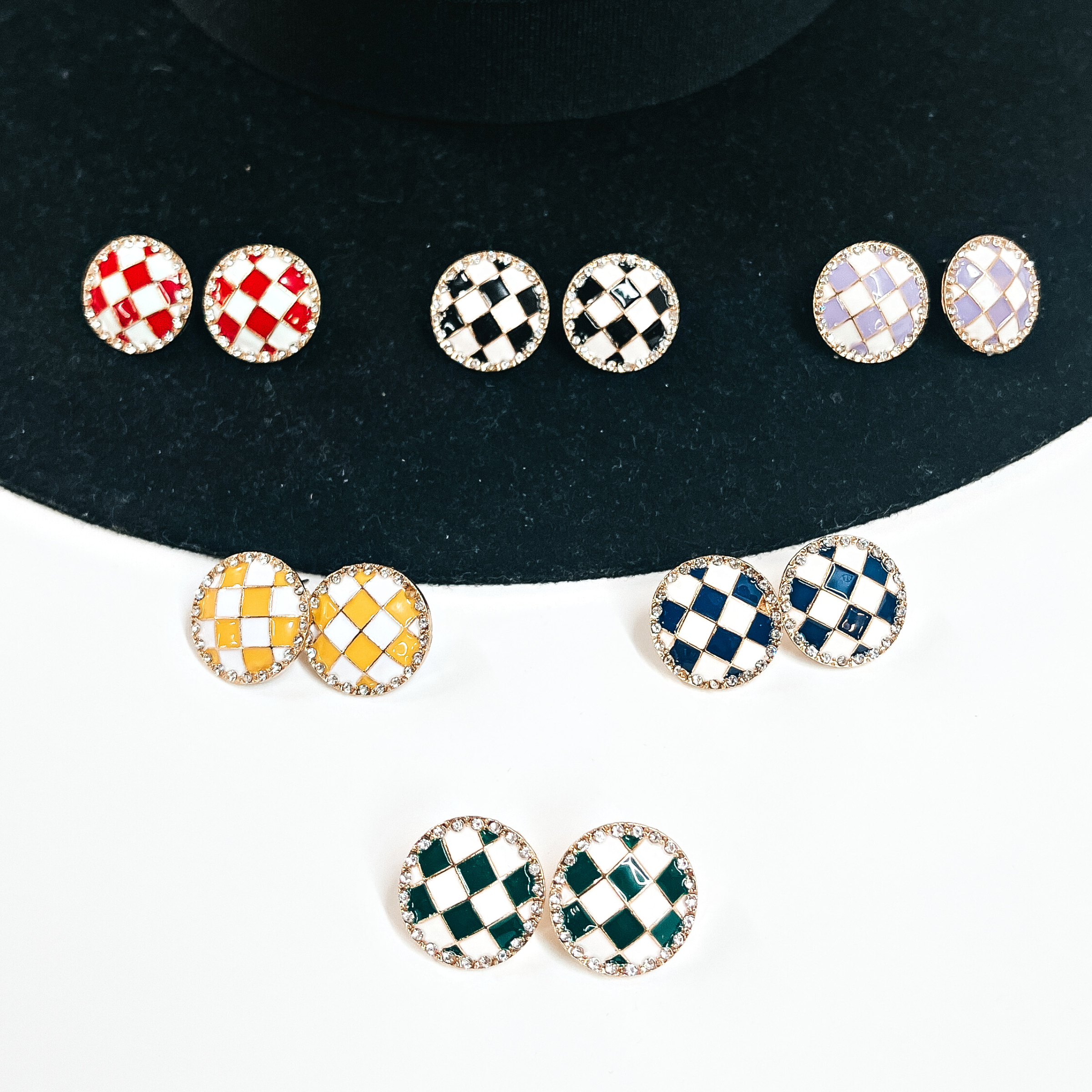 There are six pairs of checkered patterned stud earrings in different colors. The top row is red/white, black/white, and light purple/white. Middle row is yellow/white and navy blue/white, bottom pair is green/white. All of these earrings are in a gold setting with clear crystals all around. These earrings are forming a pyramid, laying on a black felt hat brim and on a white background.