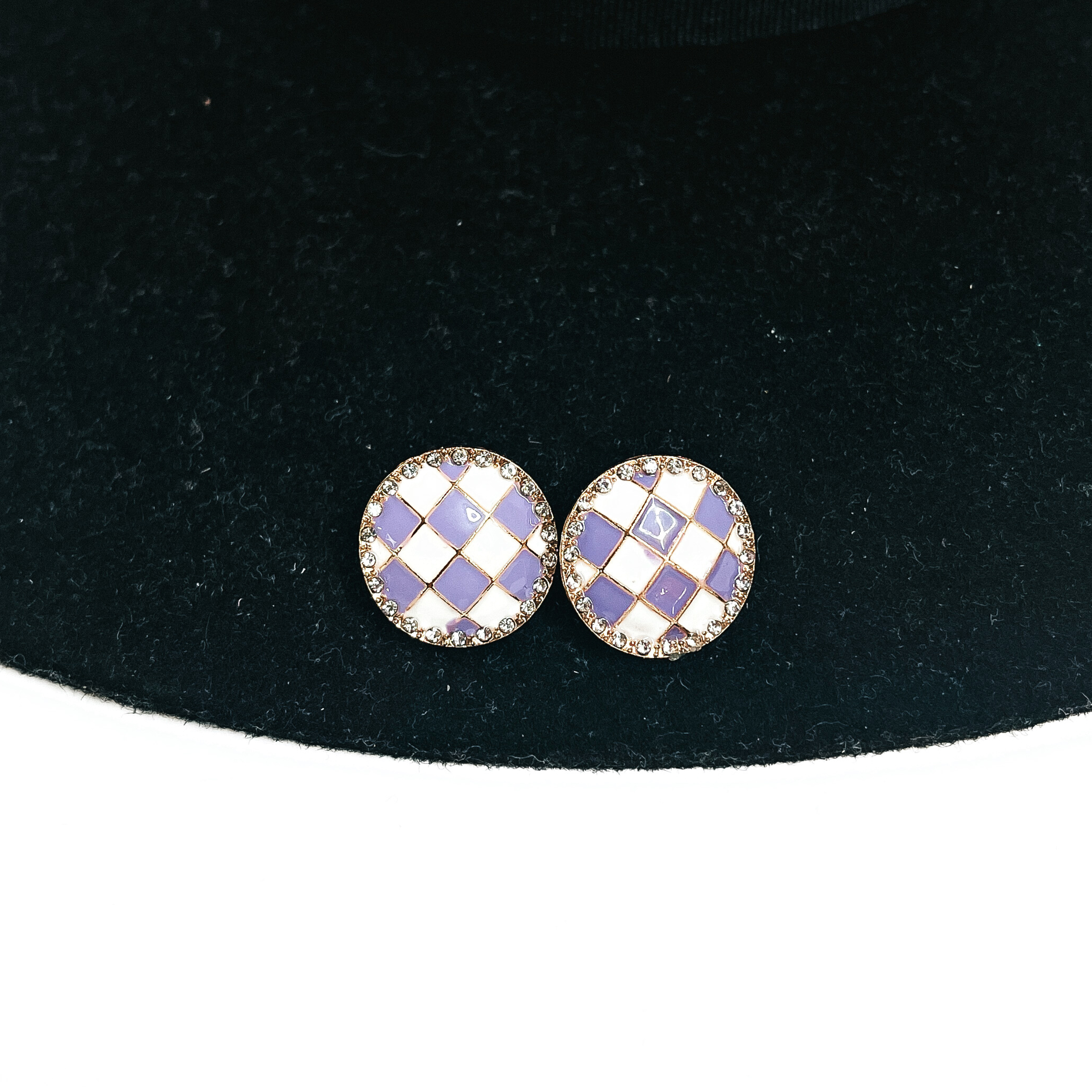 This is a pair of checkered patterned stud earrings in white/light purple in a gold  setting with clear crystals all around. This pair of earrings is laying on a  black felt hat brim and on a white background.