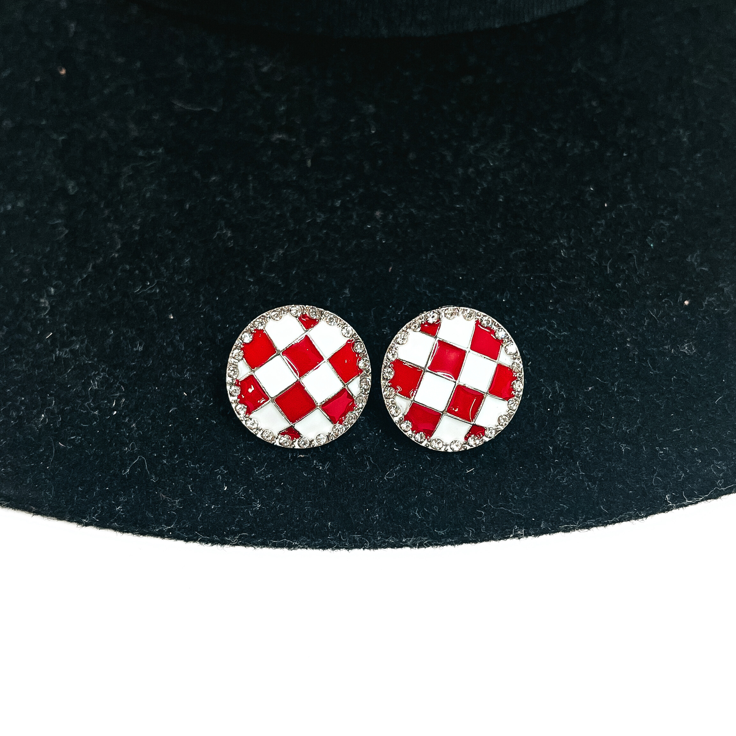 This is a white and red checkered patterned stud earrings in a silver  setting with clear crystals all around. This pair of  earrings is laying on a black felt hat brim and on a white background.