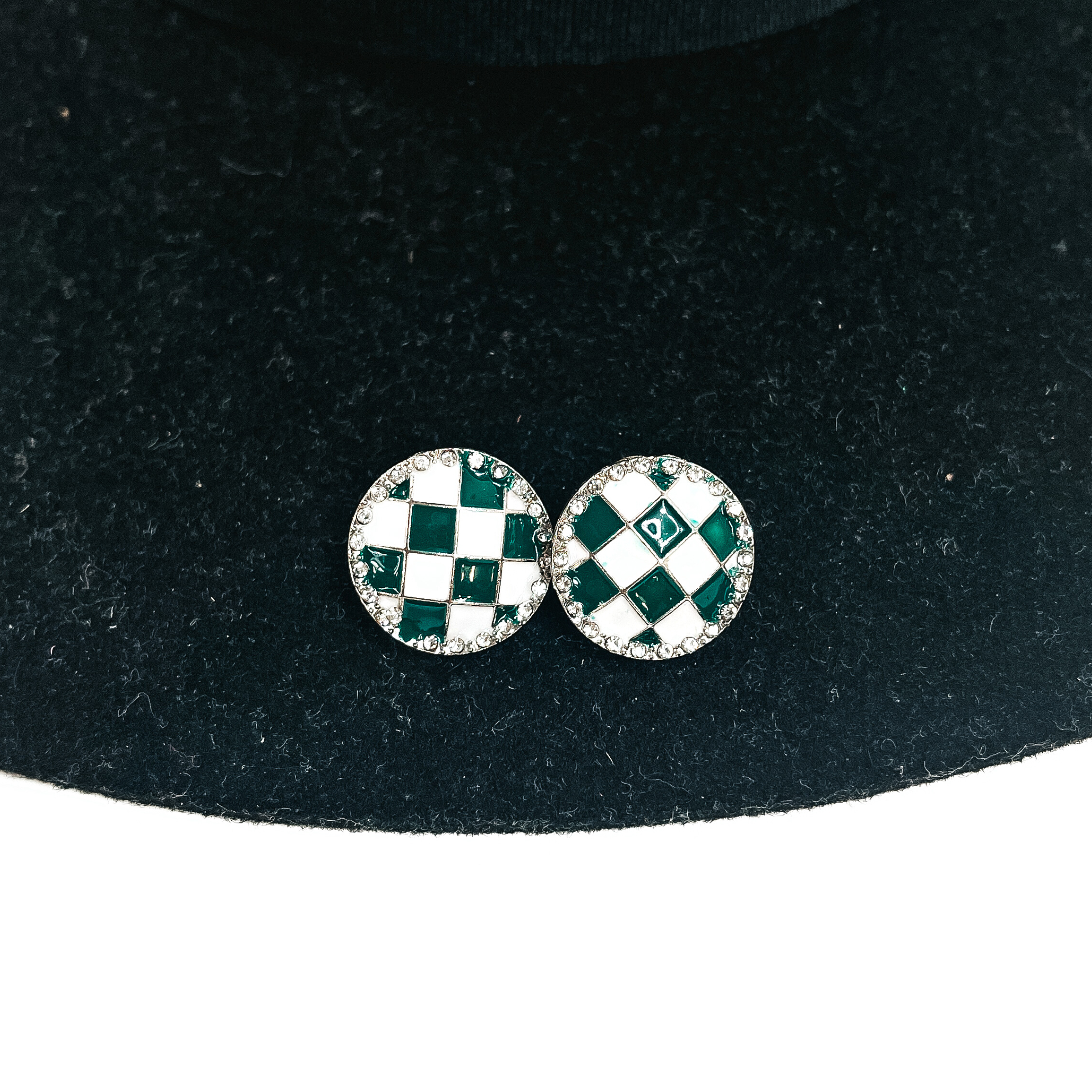 This is a white and green checkered patterned stud earrings in a silver  setting with clear crystals all around. This pair of  earrings is laying on a black felt hat brim and on a white background.