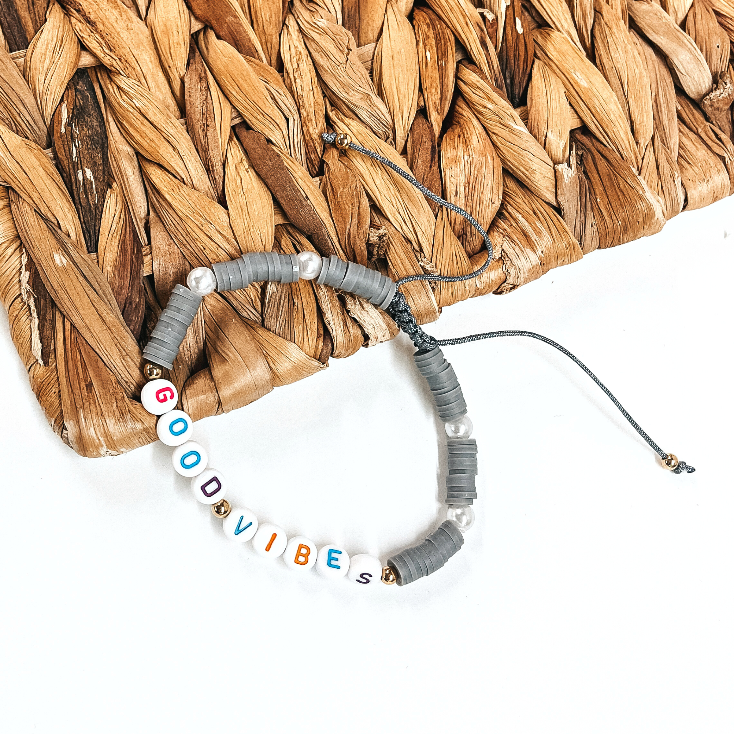This is a disk beaded bracelet in gray that says, Good Vibes in  different colors. There are pearl spacers.  This bracelet is laying partly on a woven slate and on a white background.