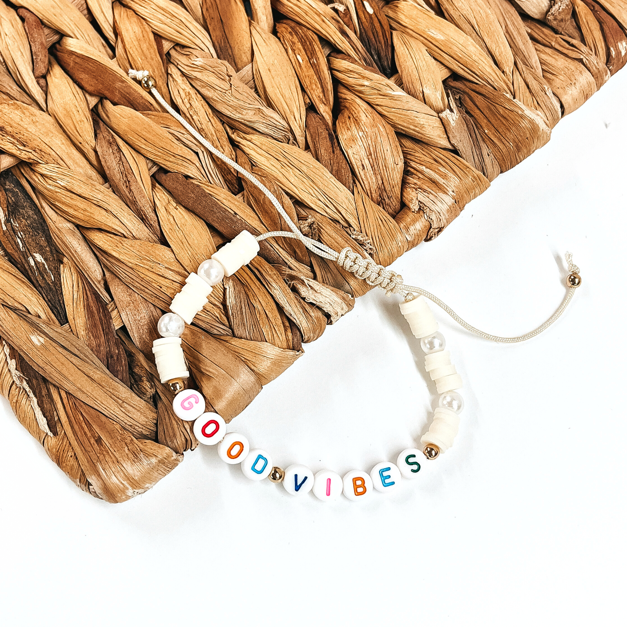 This is a disk beaded bracelet in ivory that says, Good Vibes in  different colors. There are pearl spacers.  This bracelet is laying partly on a woven slate and on a white background.