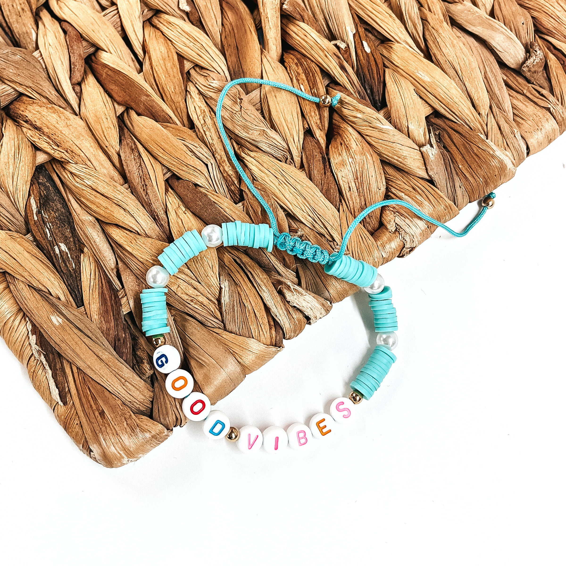 This is a disk beaded bracelet in turquoise that says, Good Vibes in different colors. There are pearl spacers. This bracelet is laying partly on a woven slate and on a white background.