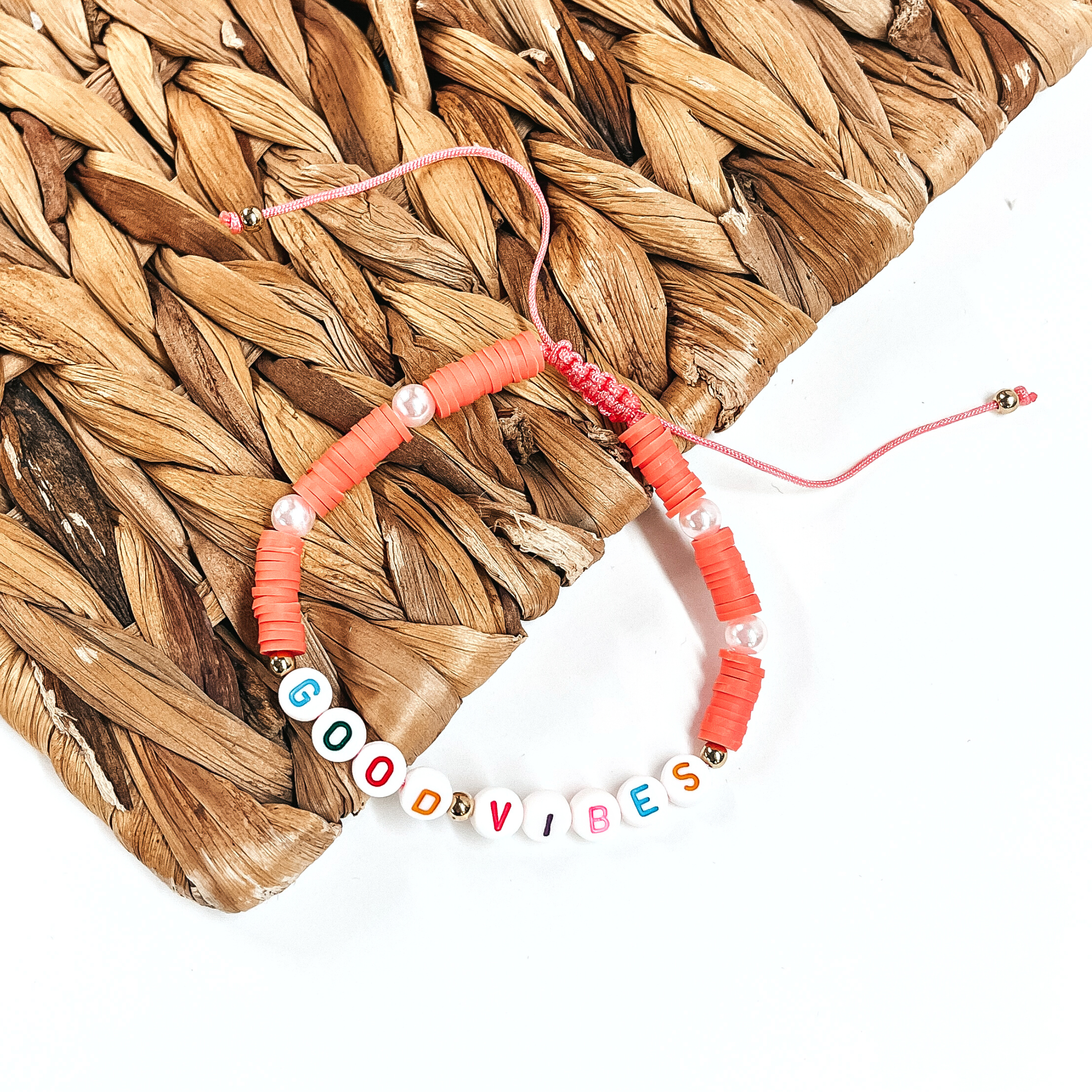 This is a disk beaded bracelet in coral that says, Good Vibes in  different colors. There are pearl spacers.  This bracelet is laying partly on a woven slate and on a white background.