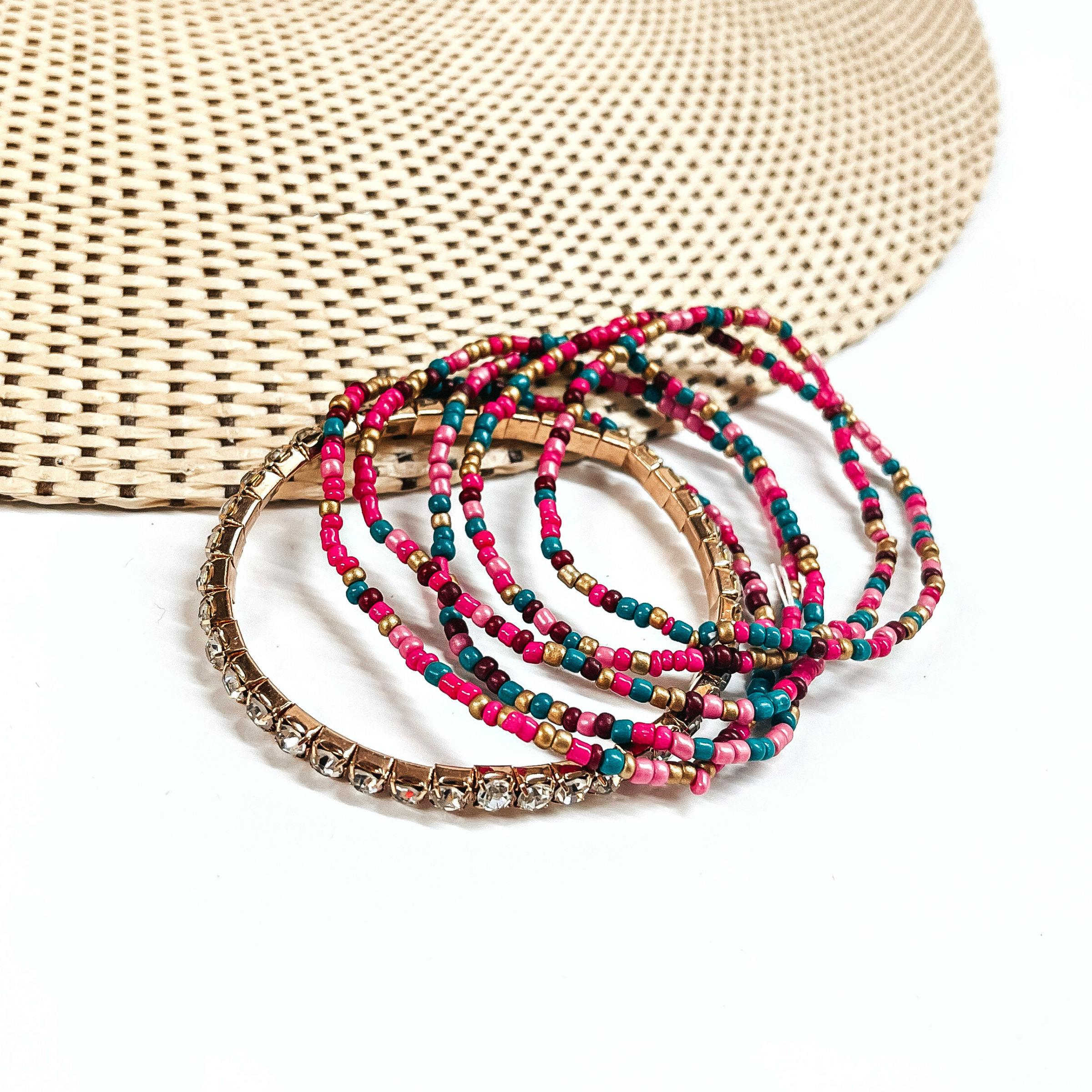 This is a set of six bracelets in a teal/pink mix  There are five seedbeaded bracelets in teal, dar purple, gold, and pink.  There is a one clear rhinestone bracelet in a gold setting.  These bracelets are laying partly on a straw hat and a white background.