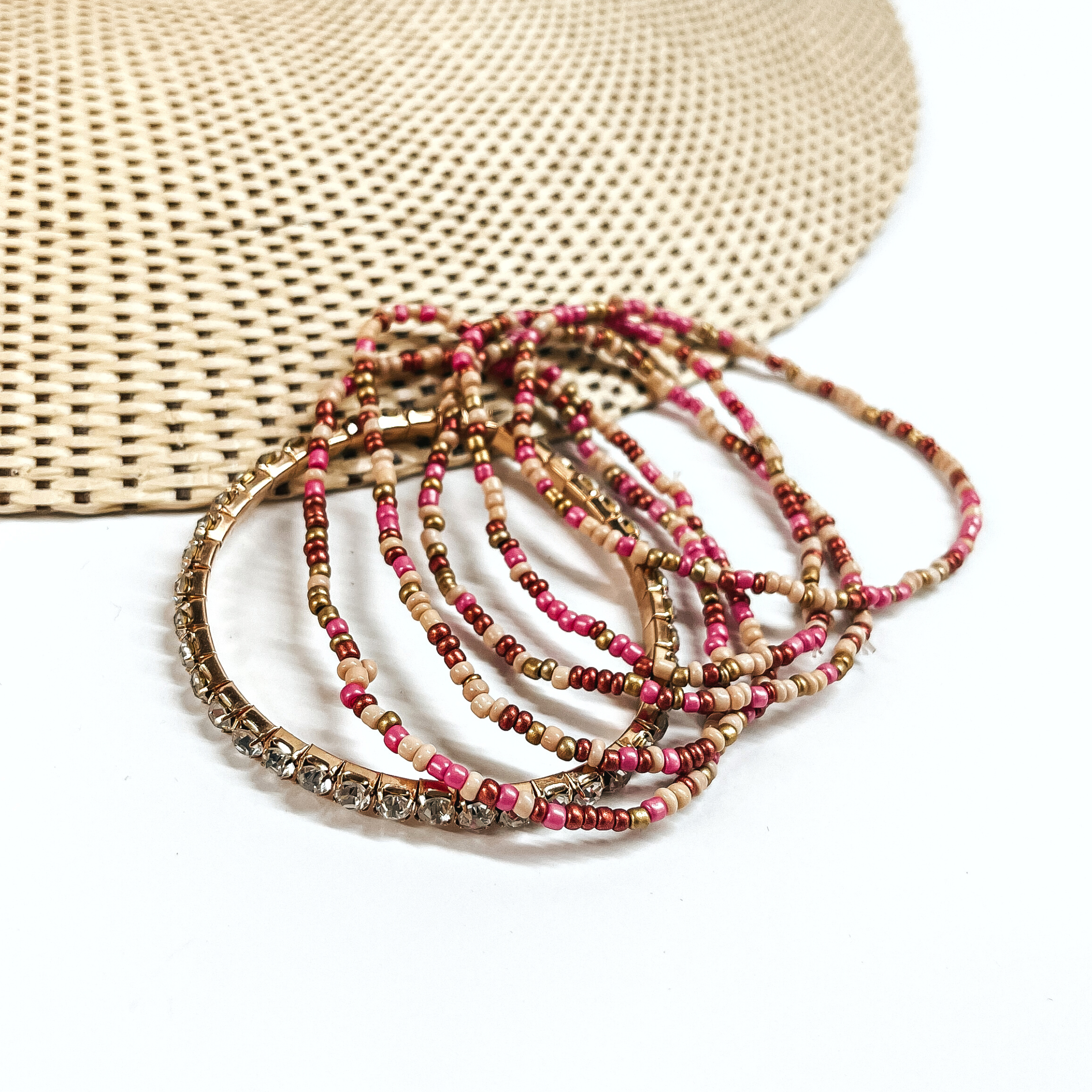This is a set of six bracelets in a gold/champagne mix  There are five seedbeaded bracelets in champagne, rose gold, gold, and pink.  There is a one clear rhinestone bracelet in a gold setting.  These bracelets are laying partly on a straw hat and a white background.