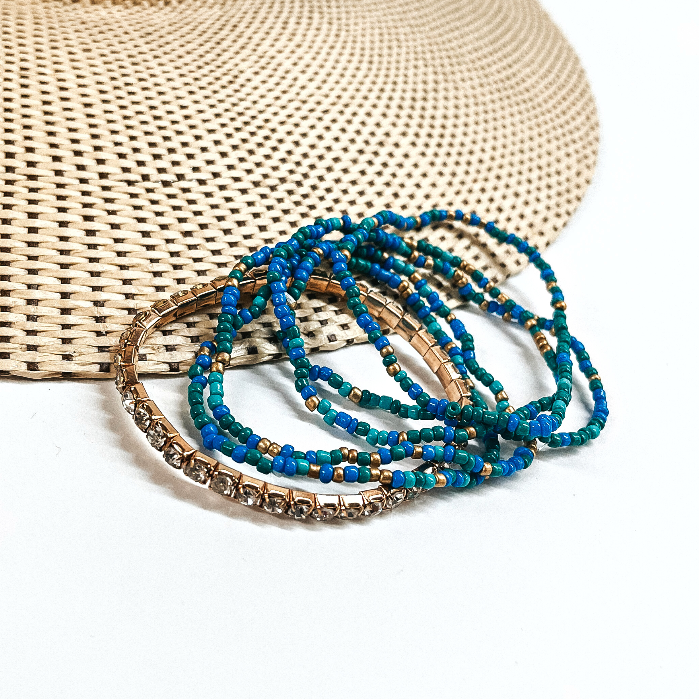 This is a set of six bracelets in a blue/teal mix. There are five seedbeaded bracelets in blue, teal, green, ad gold. There is a one clear rhinestone bracelet in a gold setting. These bracelets are laying partly on a straw hat and a white background,