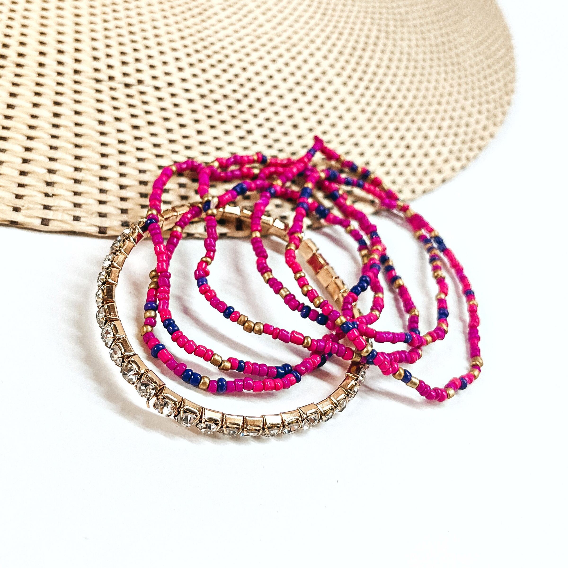 This is a set of six bracelets in a pink/purple mix  There are five seedbeaded bracelets in hot pink, purple, fuchsia, and gold.  There is a one clear rhinestone bracelet in a gold setting.  These bracelets are laying partly on a straw hat and a white background.