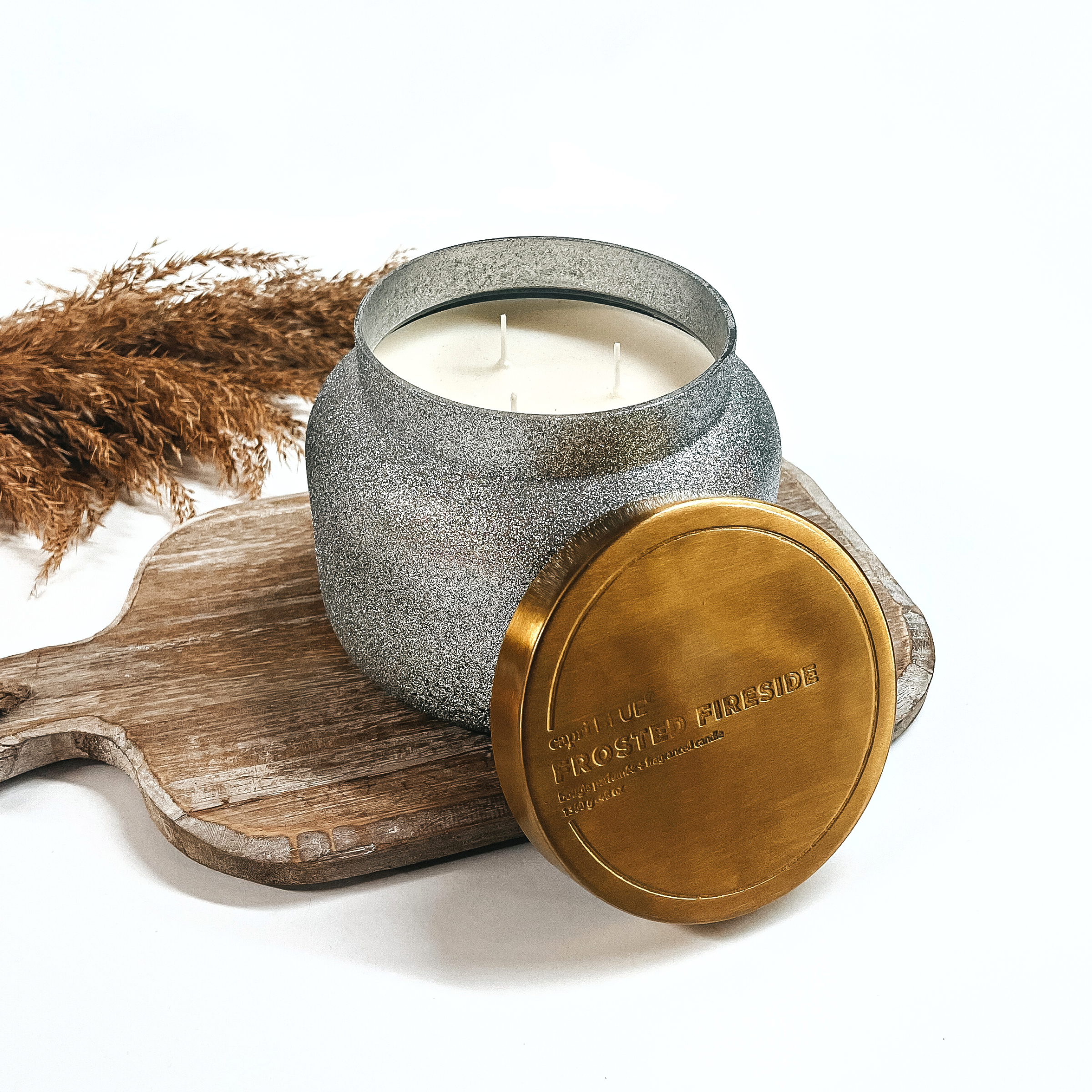 Pictured is a 48 oz. Marble Petite Jar Candle in Silver Glitter from Capri Blue. The candle scent is Frosted Fireside, which smells like smoked cedar, crackling pine and sweet peppermint. This candle has a burn time up to 100 hours. It is pictured open, with the top propped up, on a wicker tray.