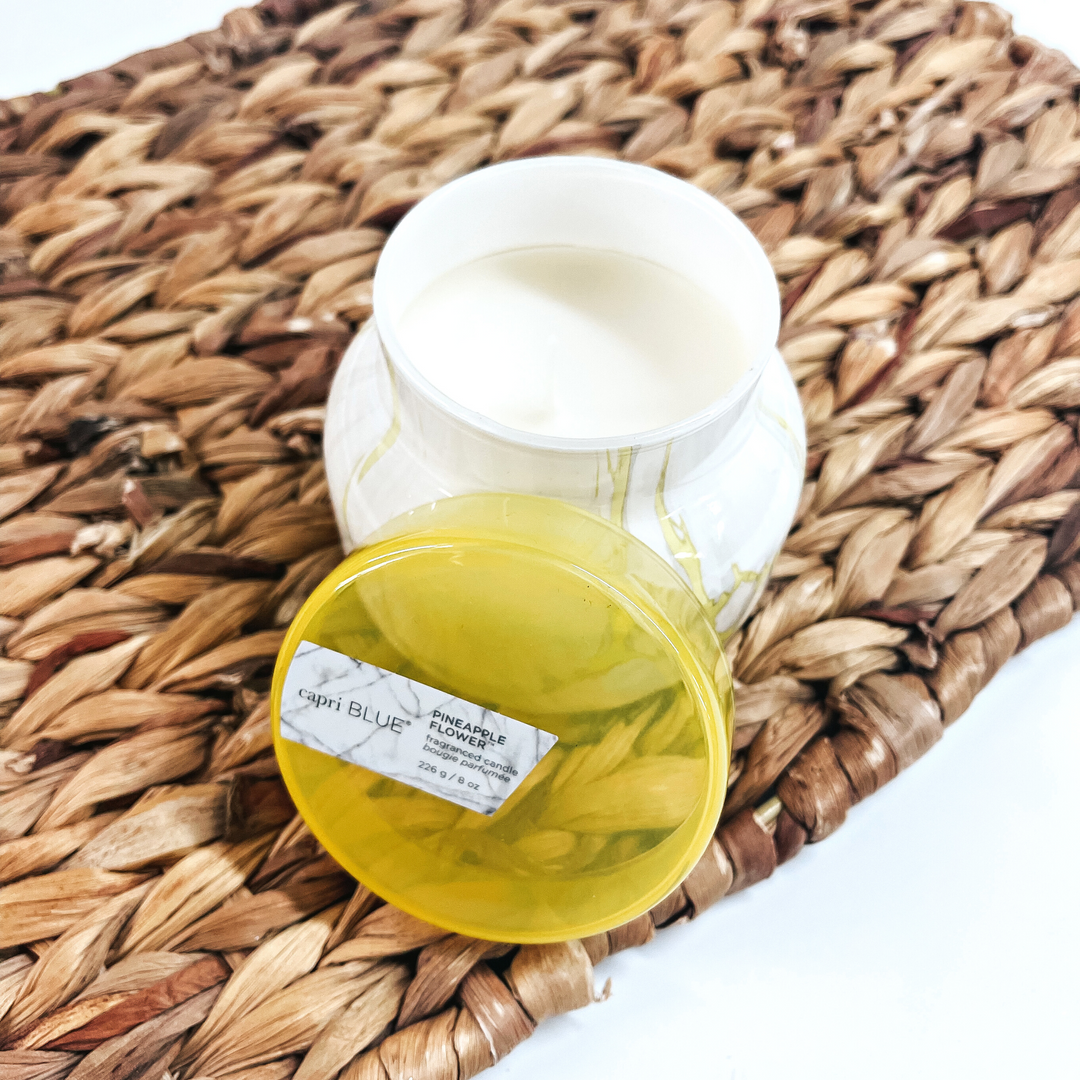 Pictured is a 8 oz. Marble Petite Jar Candle in Sunburst Yellow from Capri Blue. The candle scent is Pineapple Flower, which smells like pineapple, plum, tart lemon flower and sweet sugar cane. This candle has a burn time up to 40 hours. It is pictured open, with the top propped up, on a wicker tray.