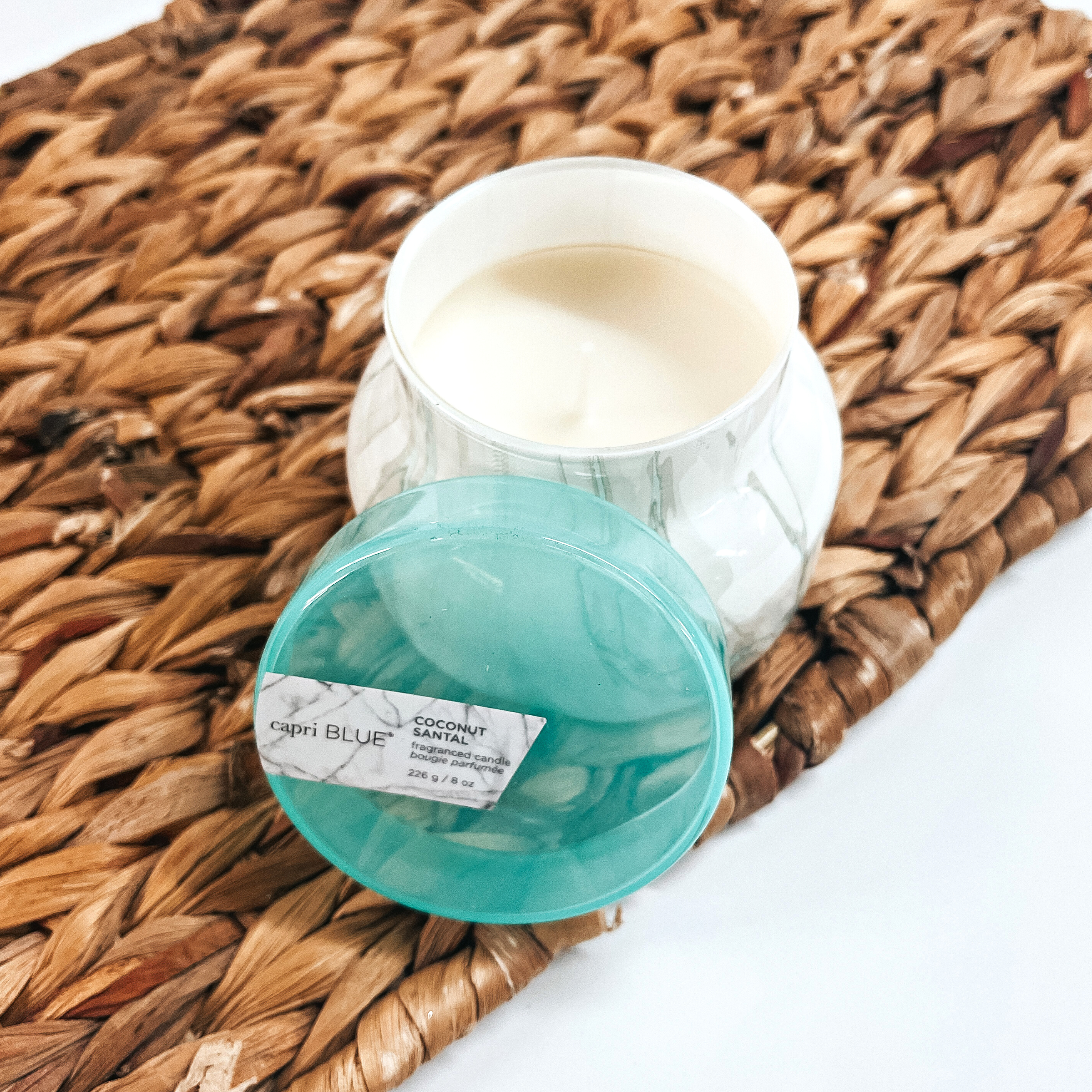 Pictured is a 8 oz. Marble Petite Jar Candle in Sea Glass from Capri Blue. The candle scent is Coconut Santal, which smells like hibiscus, lime coconut and hits of amber. This candle has a burn time of up to 40 hours. It is pictured open, with the top propped up, on a wicker tray.