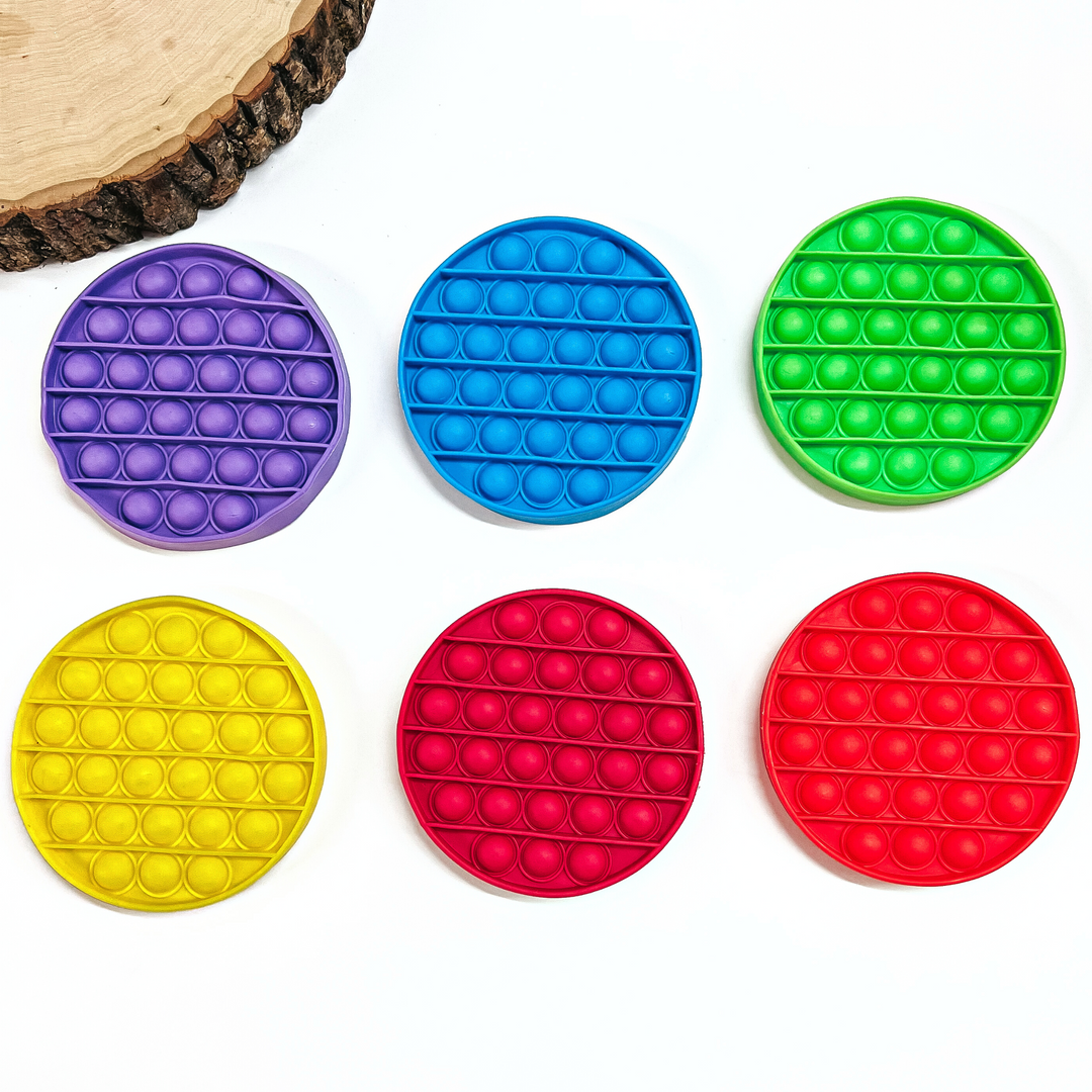 There are six, rubber, circle pop toys in various colors. Purple, blue, green, yellow, magenta, and red. These toys are laying on a white background with a slab of wood in the side as decor.