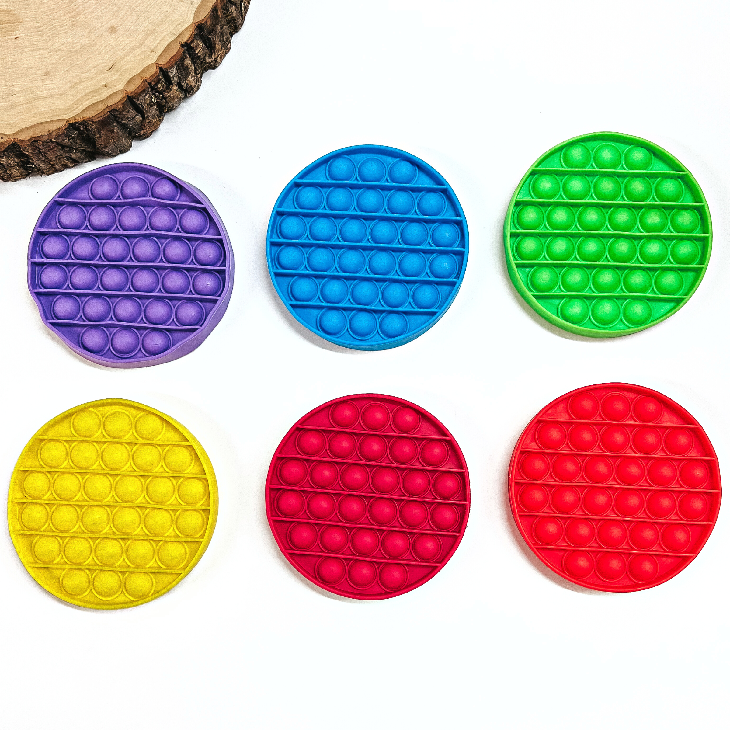 There are six, rubber, circle pop toys in various colors. Purple, blue, green, yellow, magenta, and red. These toys are laying on a white background with a slab of wood in the side as decor.