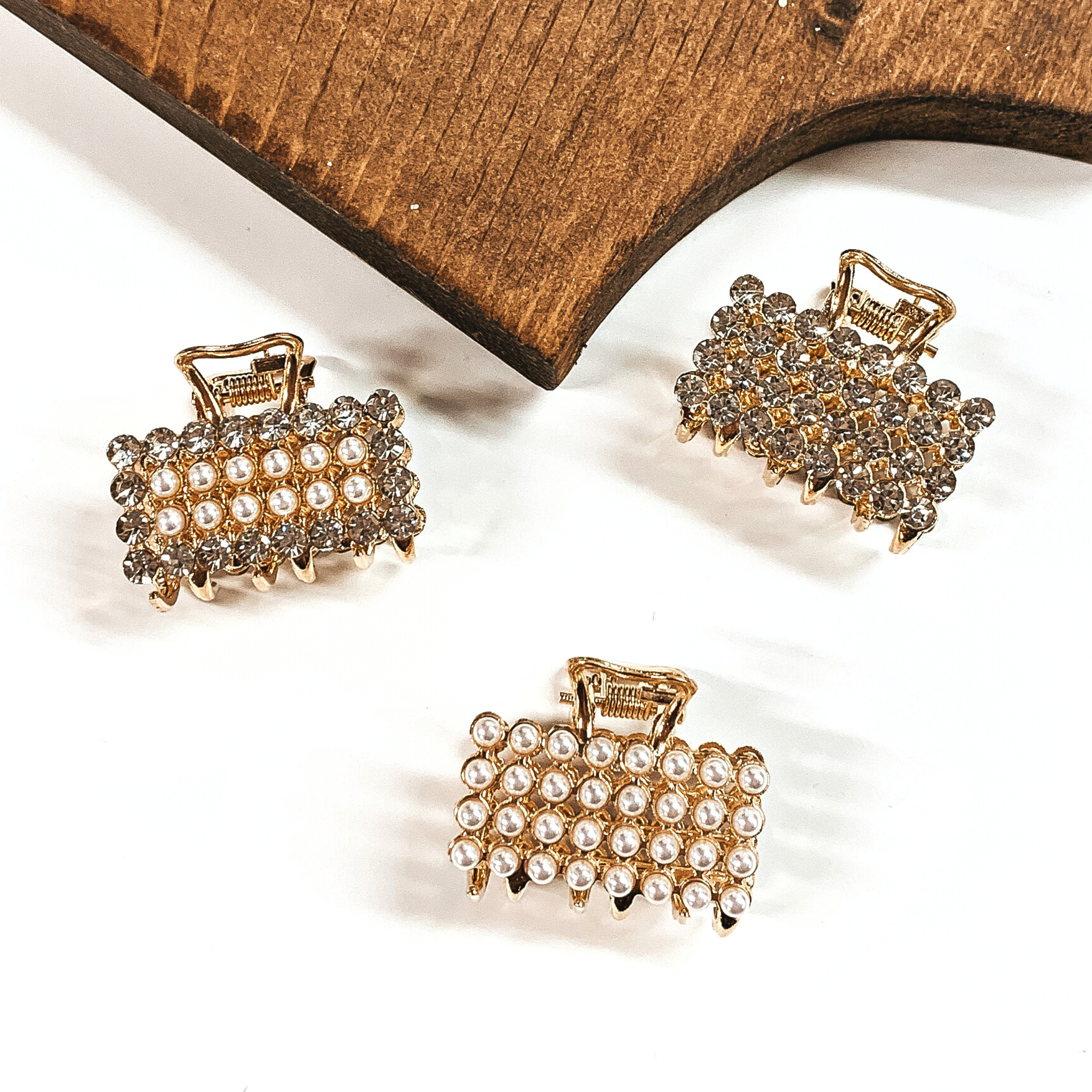 There are three small rectangle clips in gold with different materials. There is a clear crystal and pearl one, clear crystals, and all pearls. These clips are taken on a white background with a slab of wood in the back as decor.
