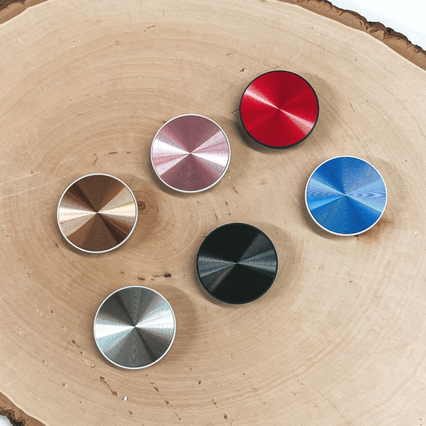 There are six circle phone grips in metallic colors with a swil texture. There is copper, pink, red, silver, black, and blue. These phone grips are taken on a slab of wood .