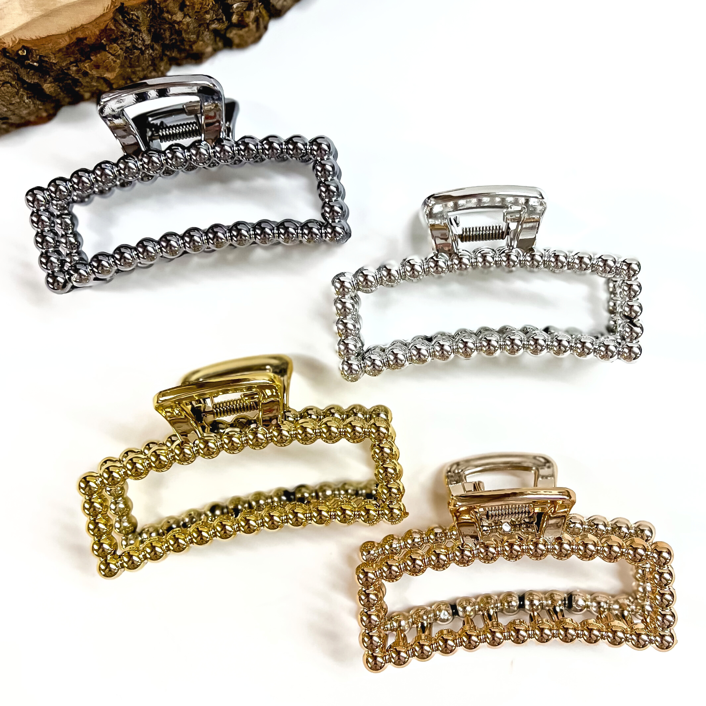There are four rectangle metallic clips in different colors, all clips have a beaded texture all over them. There is gunmetal, silver, gold, and copper. These clips are laying on a white background with a slab of wood in the top as decor.