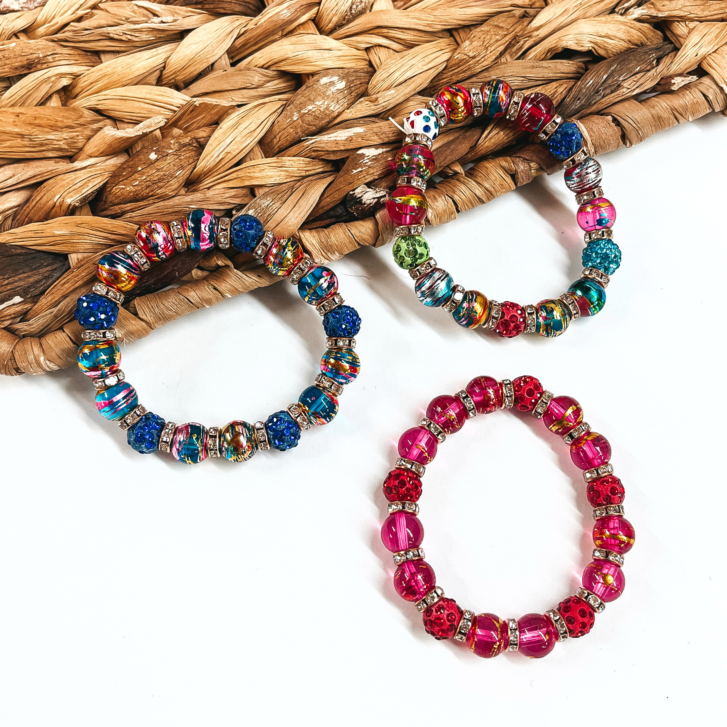There are three multicolored beaded bracelets with crystals. the left one is a blue mix with crystals, the right one is multicolored mix with crystals, and the bottom one is red/pink mix with crystals. All three are laying on a brown woven slate and a white background.