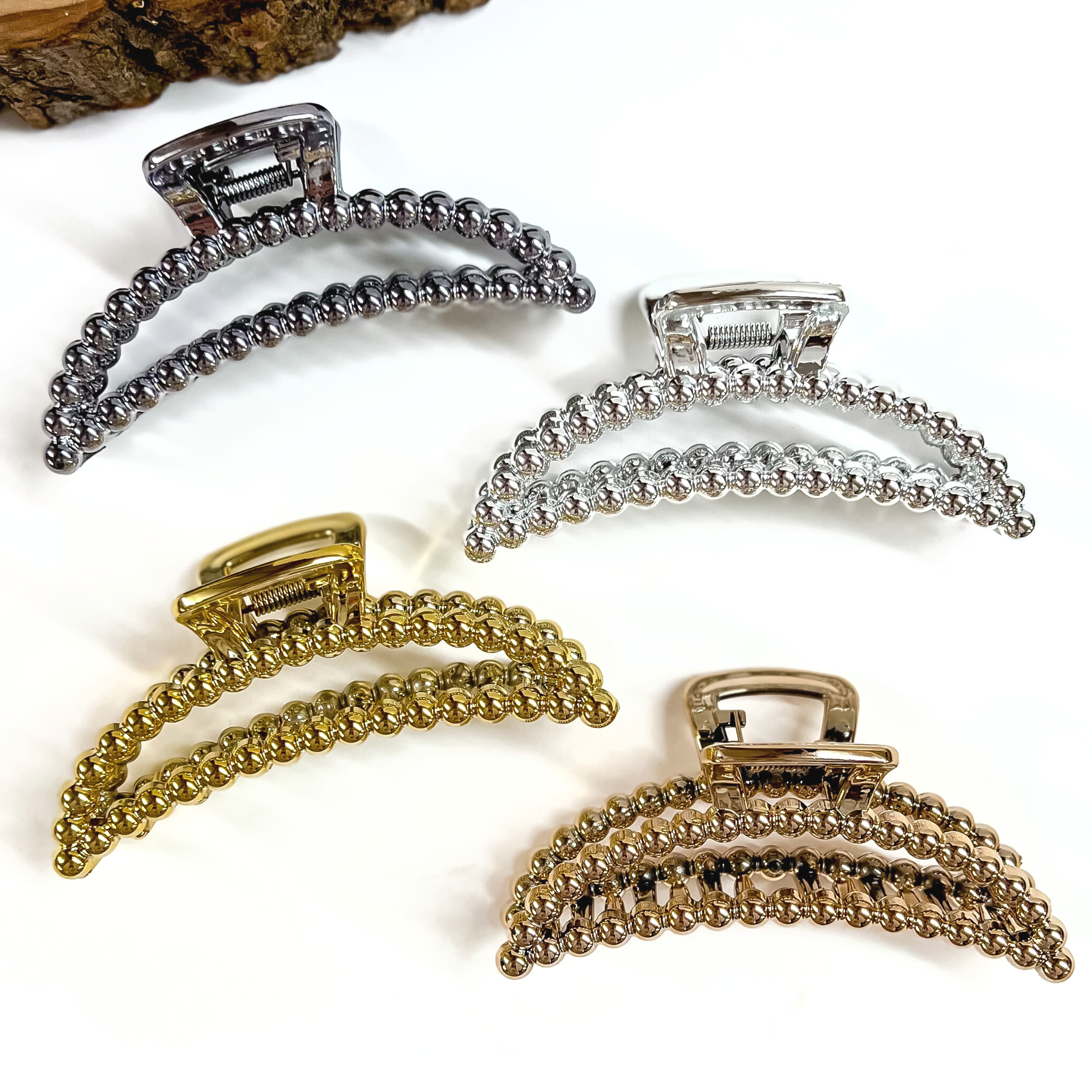 There are four metallic curved clips on different colors. They all have beaded texture all around, there is gunmetal, silver, gold, and copper. These clips are laying on  a white background with a slab of wood in the top as decor.