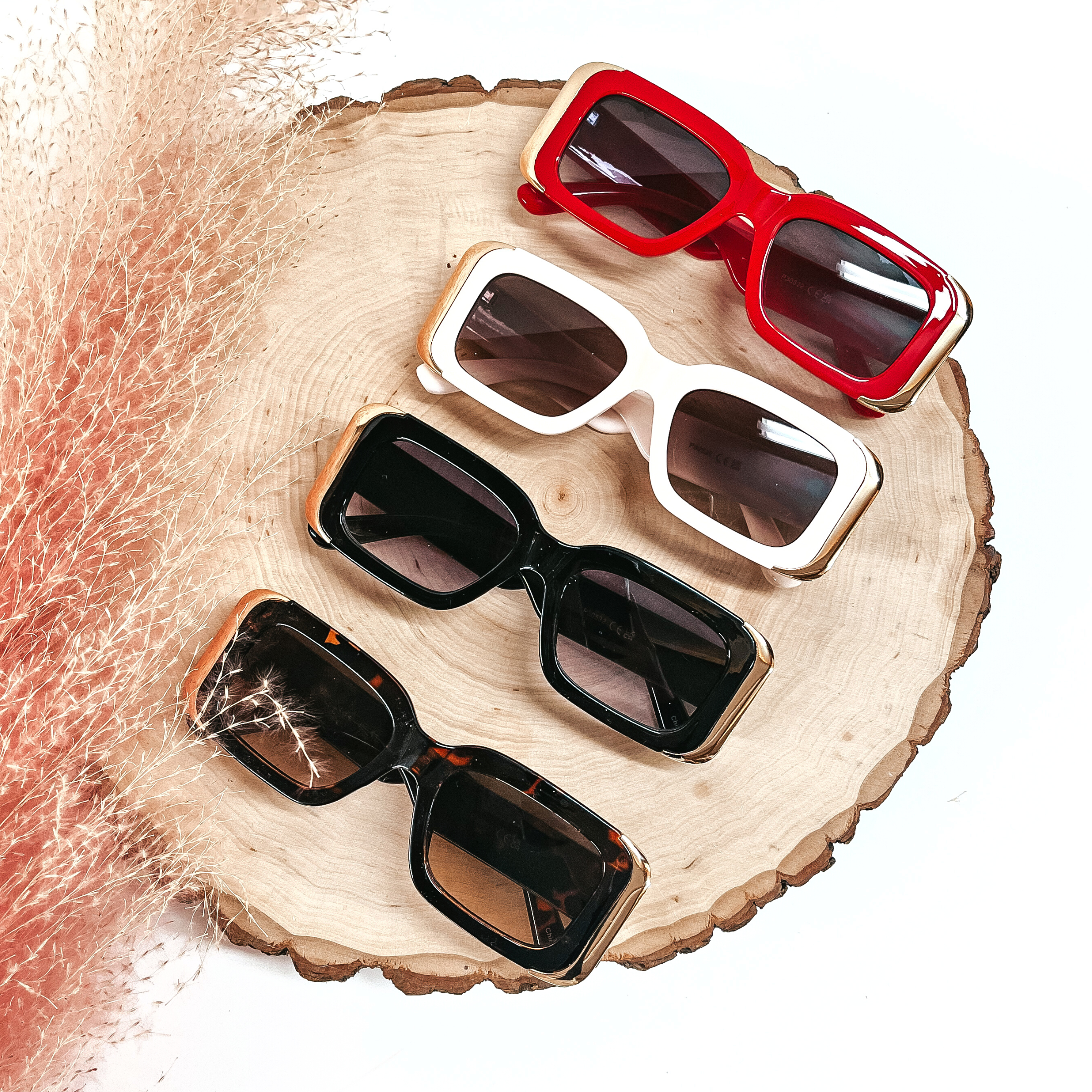 There are four rectangle sunglasses in different colors laying on a slab of wood with a pink plant in the side as decor. From top to bottom; red frames/brown lenses/gold detailing, ivory frames/brown lenses/gold detailing, black frame/black lenses/gold detailing, tortoise print frame/brown lenses/gold detailing. 