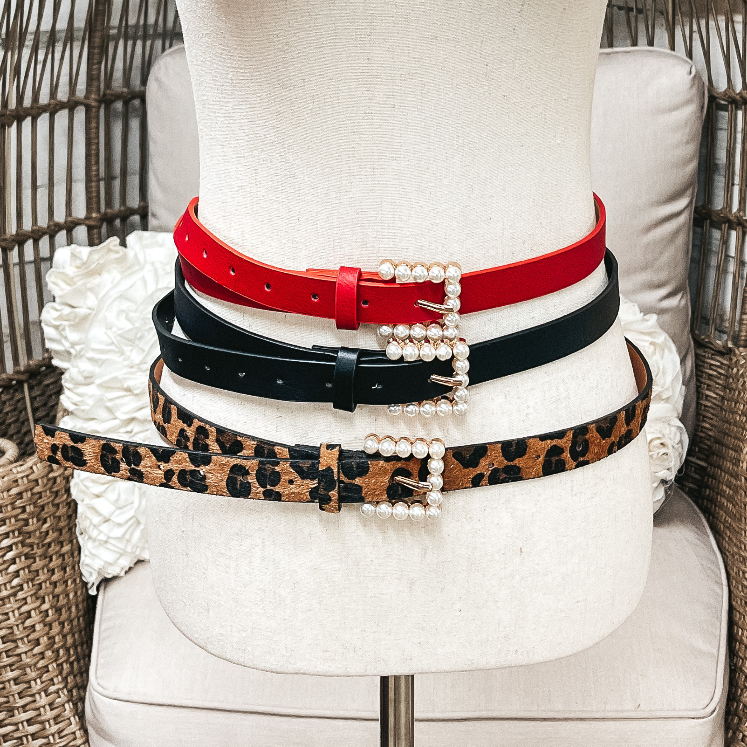 There are three skinny belts in three different colors on an ivory mannequin. From top to bottom; red, black, and leopard print, all three belts have a small rectangle pearl embellished buckle.