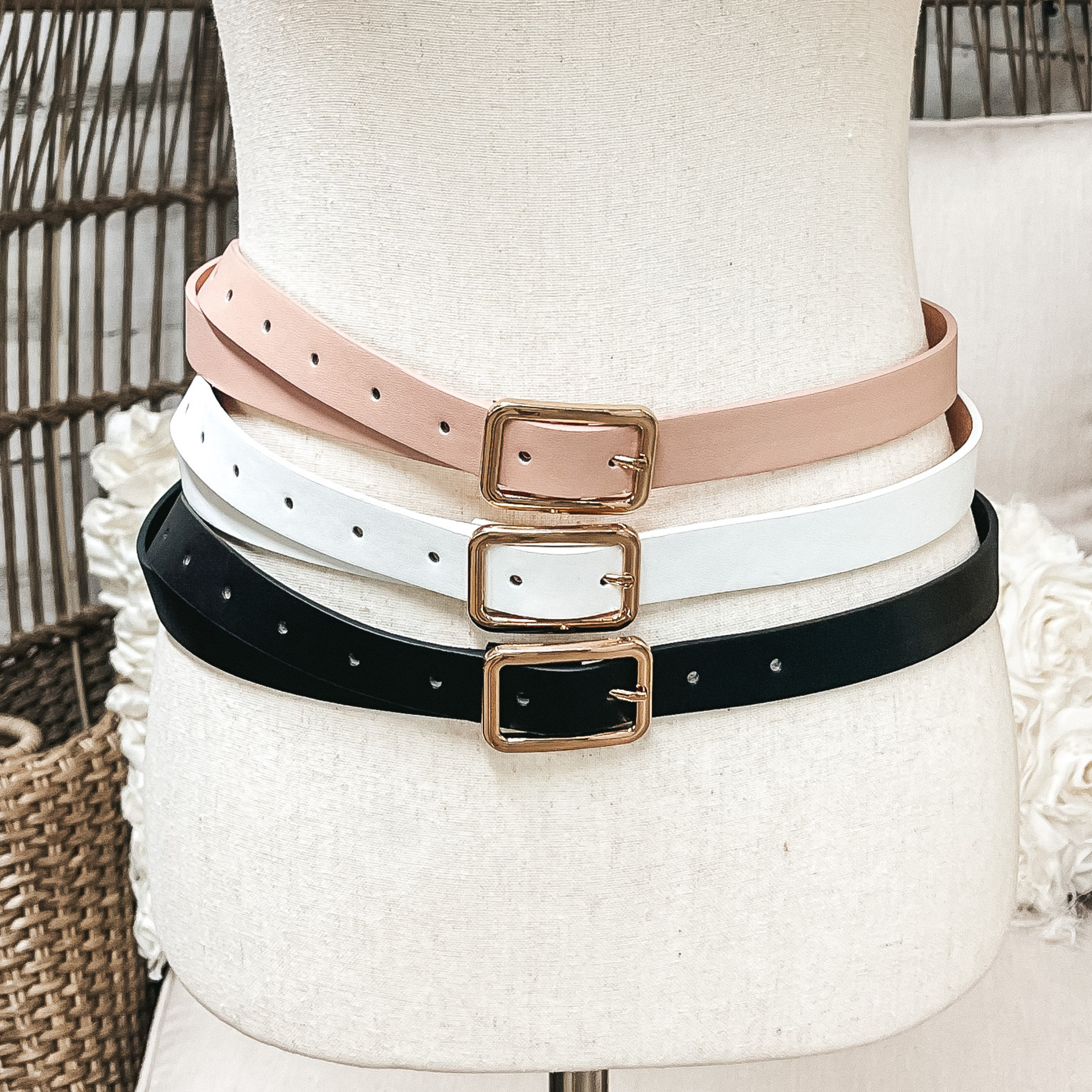 There are three skinny belts in three different colors on an ivory mannequin. From top to bottom; nude, white, and black, all three belts have a gold rectangle buckle.