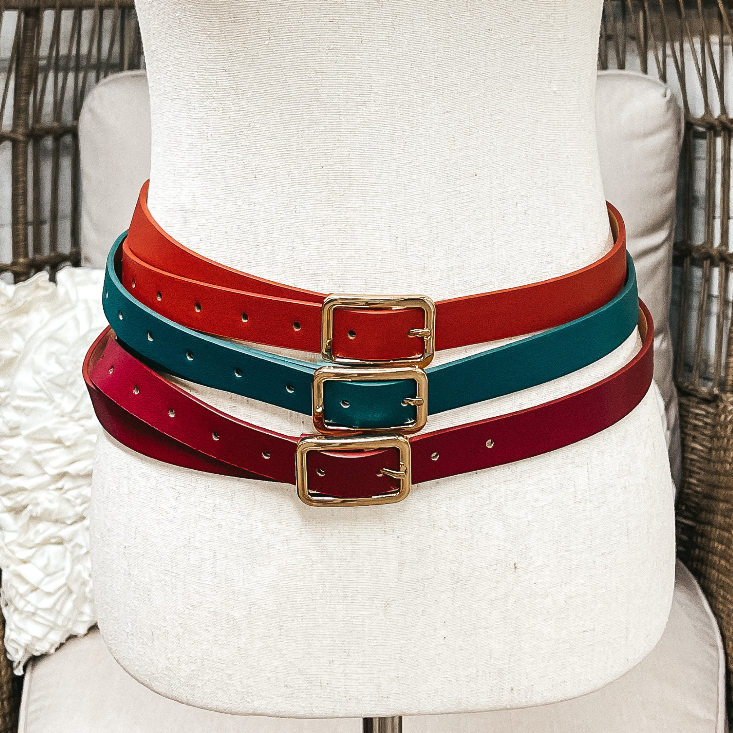 There are three skinny belts in three different colors on an ivory mannequin. From top to bottom; cognac, dark teal/green, and burgundy, all three belts have a gold rectangle buckle.
