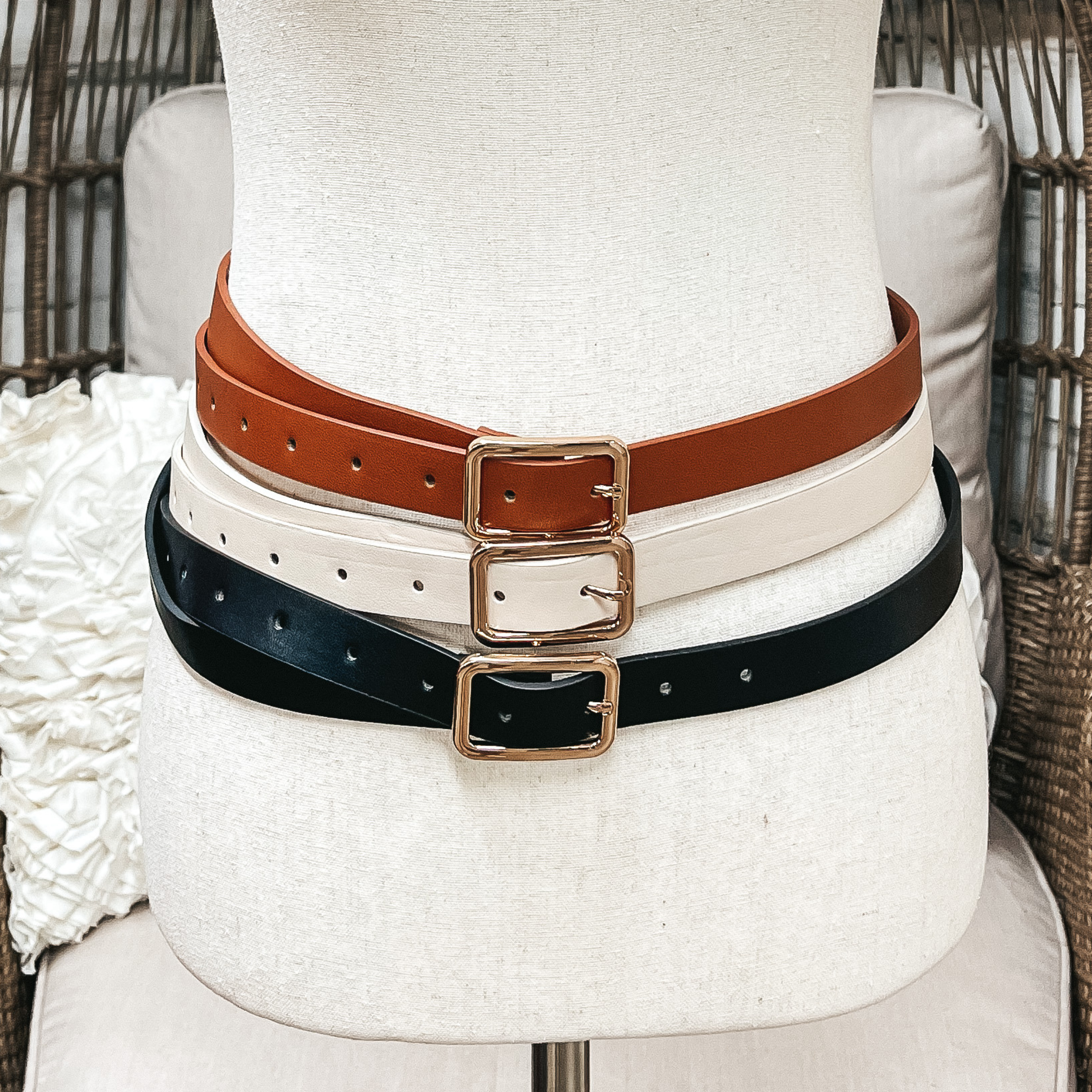 There are three skinny belts in three different colors on an ivory mannequin. From top to bottom; cognac, ivory, and black, all three belts have a gold rectangle buckle.
