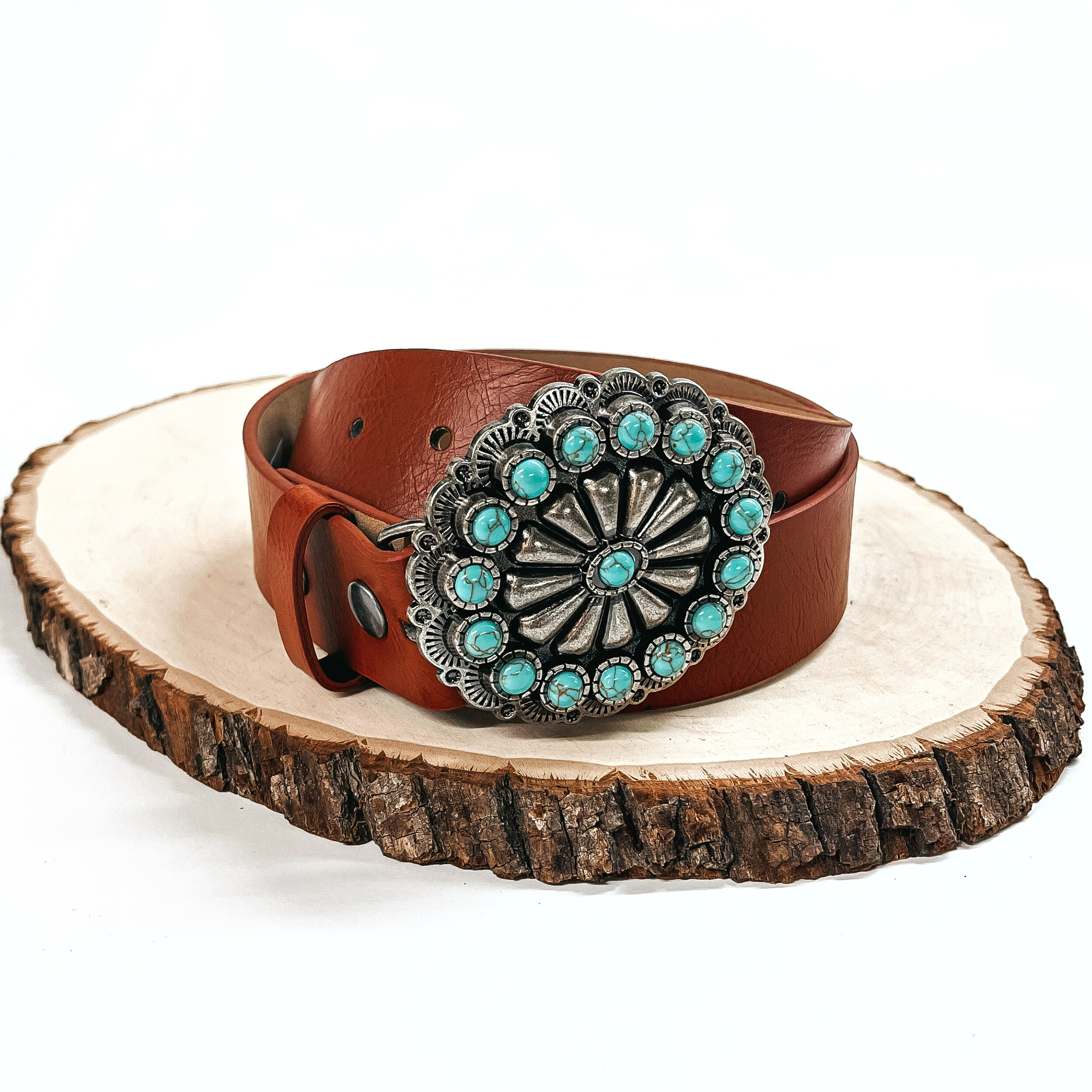 There is a brown belt with a silver concho and small turquoise stones all  around. The belt is rolled up and placed on a slab of wood on a white  background.