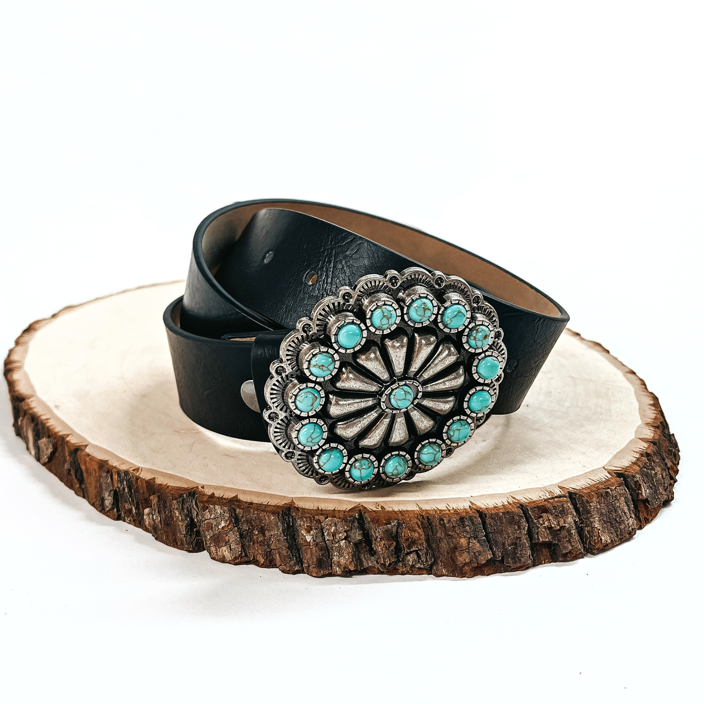 There is a black belt with a silver concho and small turquoise stones all  around. The belt is rolled up and placed on a slab of wood on a white  background.