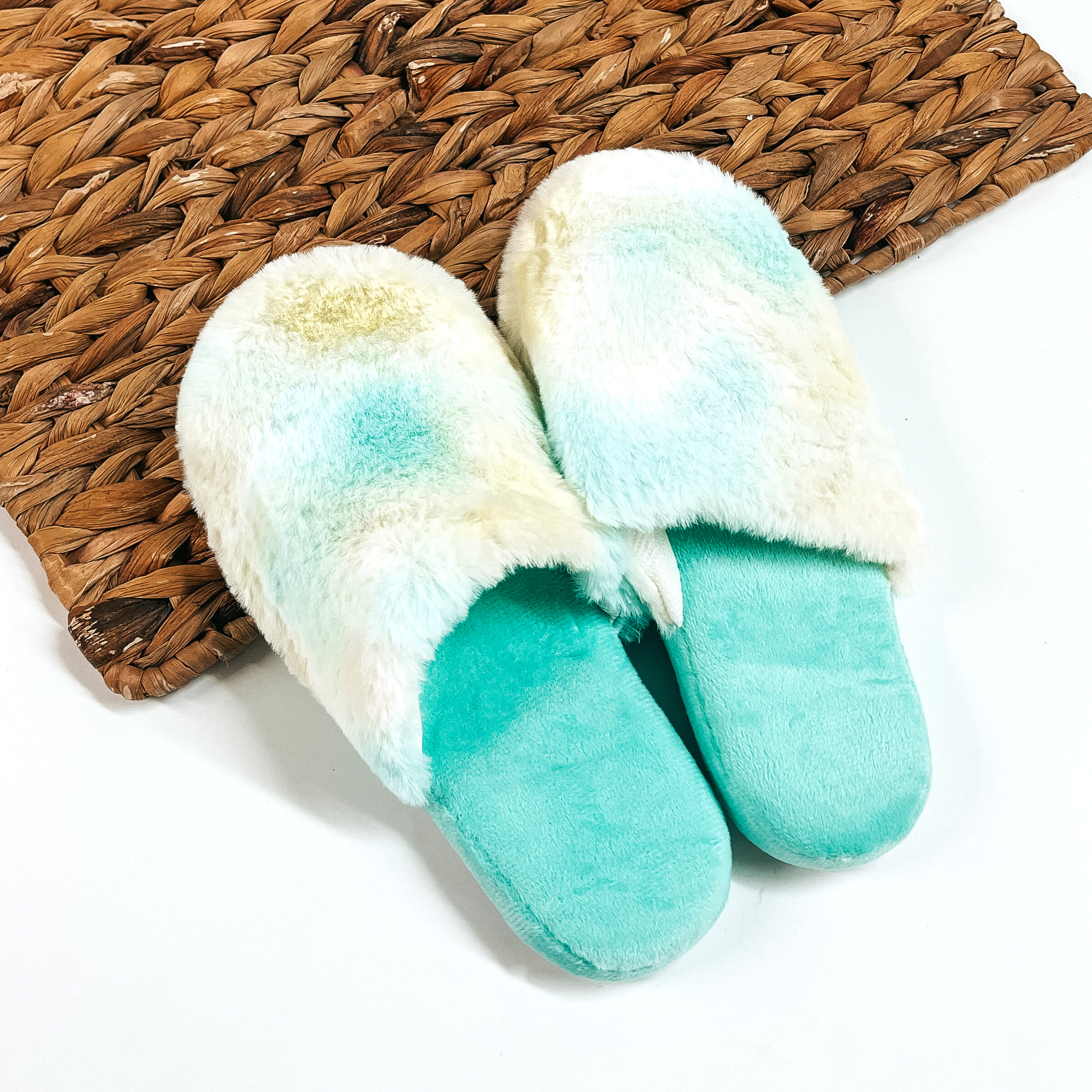There are a pair of fluffy slip on slippers, the sole is teal and the top  part is tie dye with a teal/yellow/green mix. These slippers are placed on a  white background and a brown woven slate.