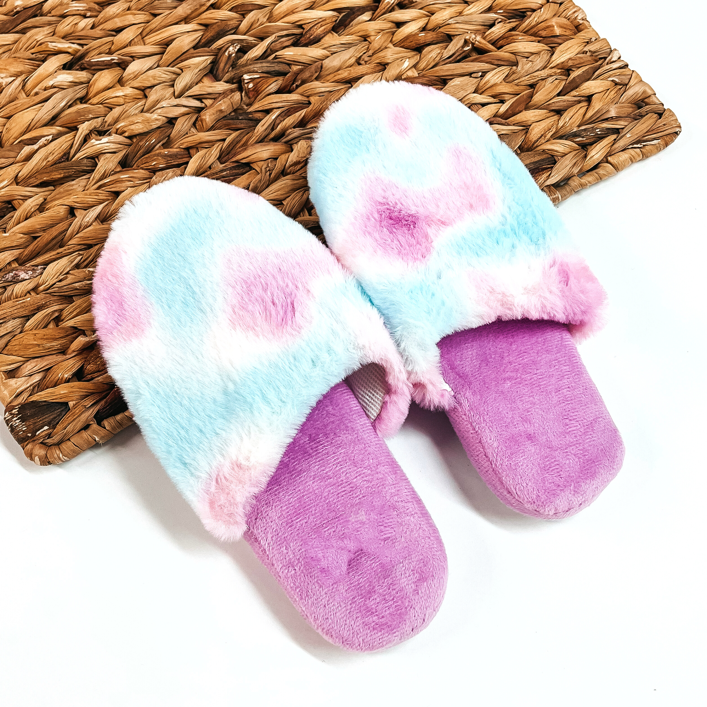 There are a pair of fluffy slip on slippers, the sole is purple and the top  part is tie dye with a purple/white/blue mix. These slippers are placed on a  white background and a brown woven slate.