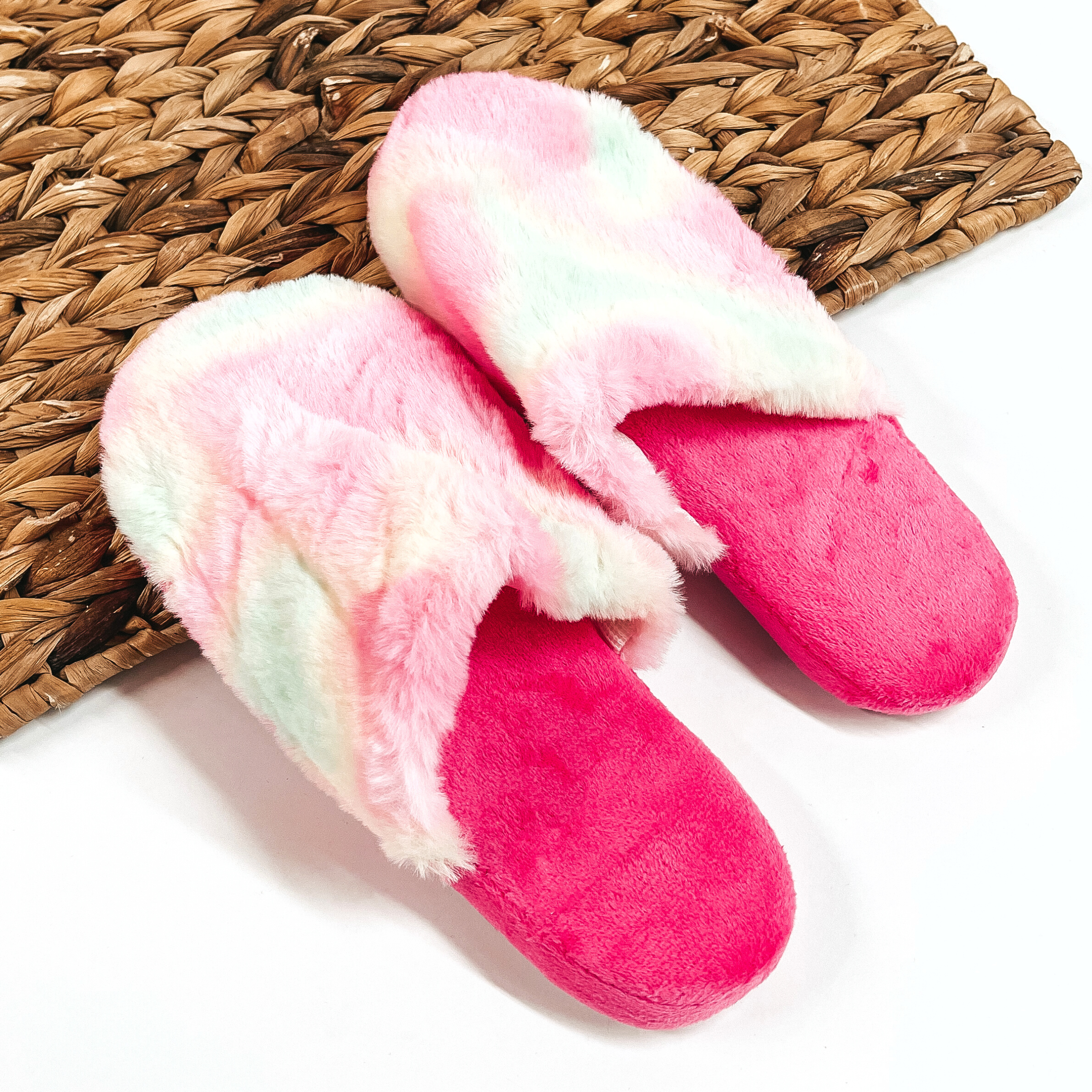 There are a pair of fluffy slip on slippers, the sole is hot pink and the top  part is tie dye with a pink/yellow/green mix. These slippers are placed on a  white background and a brown woven slate.