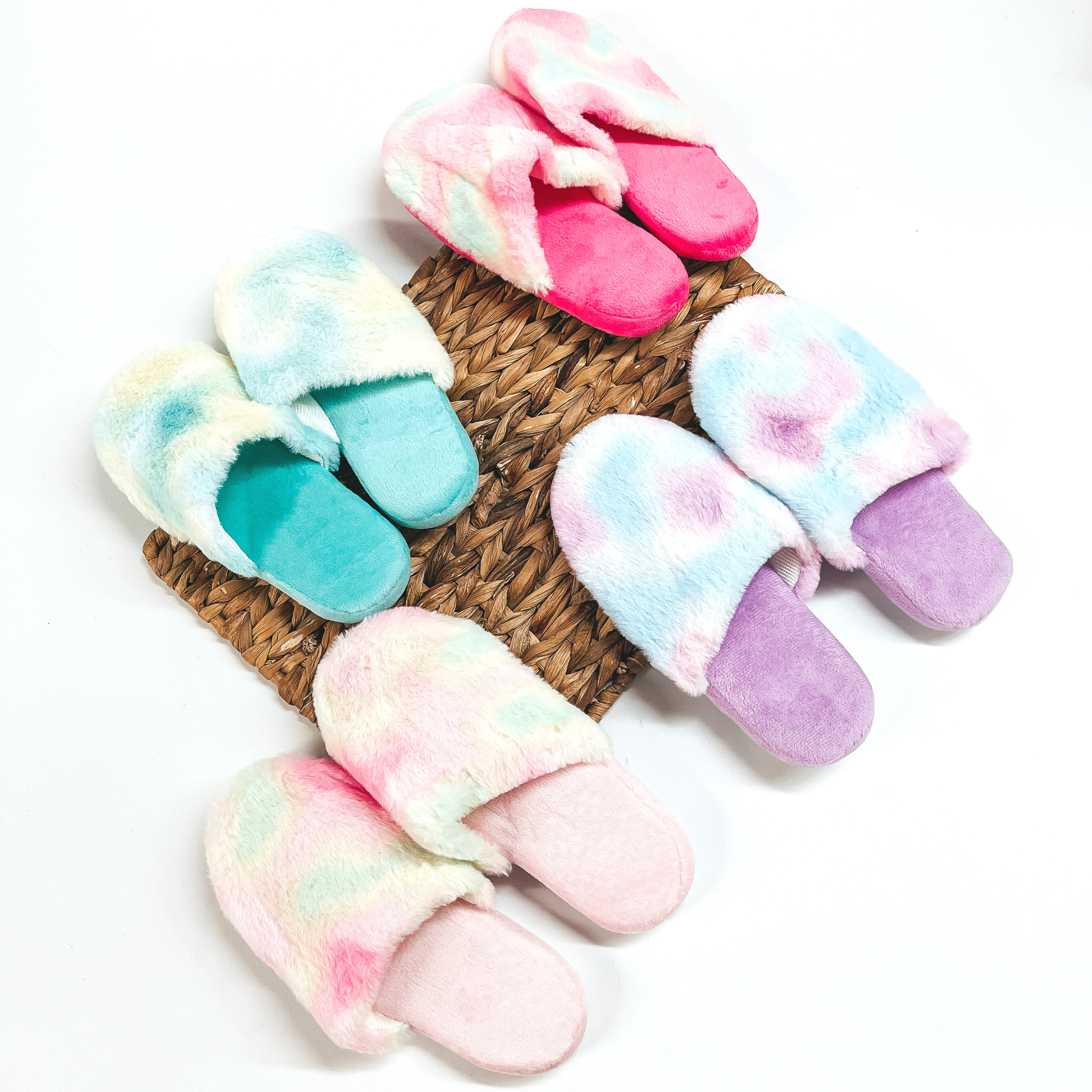 There are four pairs of fluffy tie dye slippers in hot pink, teal, purple,  and light pink. All slippers are placed on a brown woven slate and on a white  background.