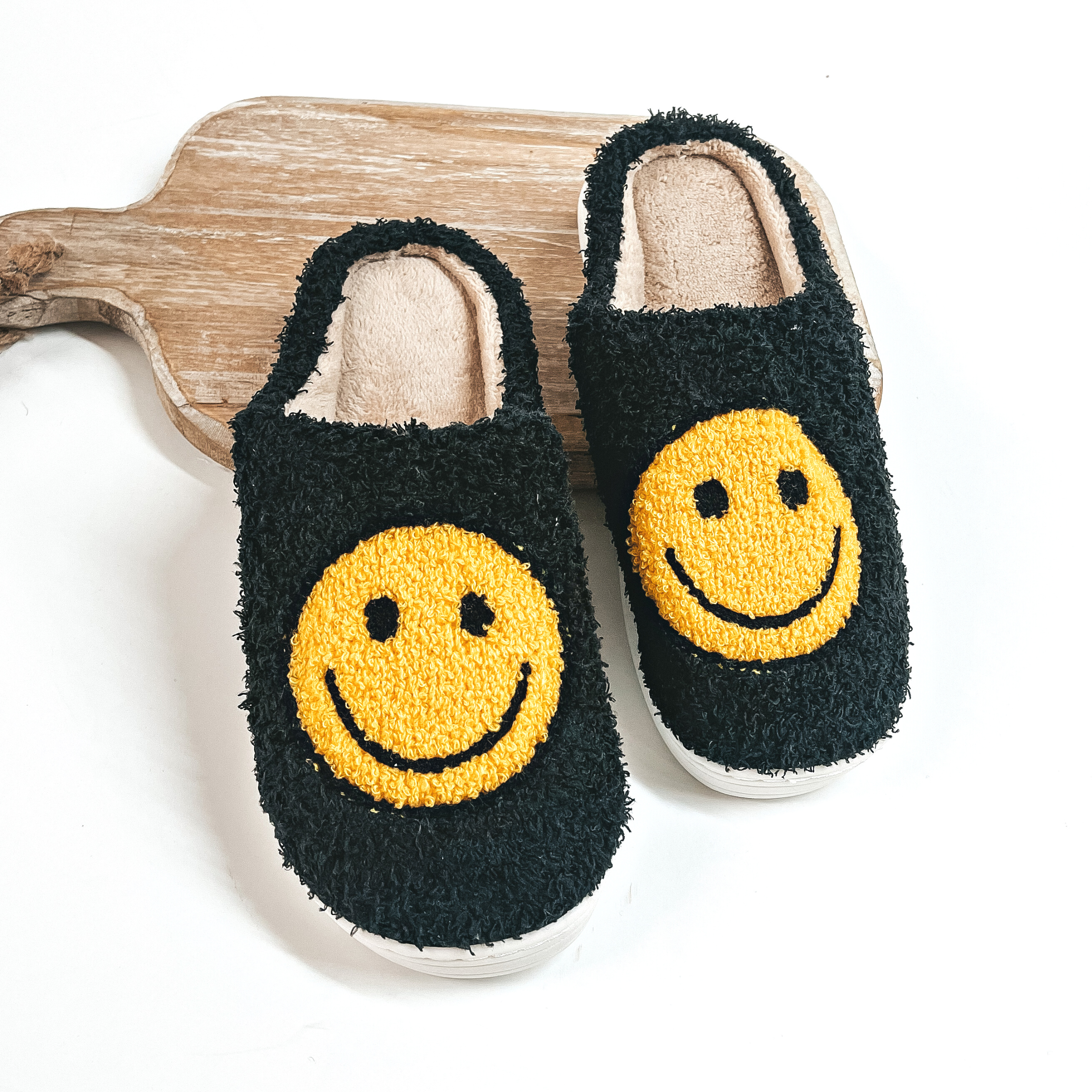 There are sherpa slippers in black with yellow happy faces on top of each  slipper. The inside is in brown and the bottom sole is white. The pair  of slippers are placed on a wooden slab and on a white backround.