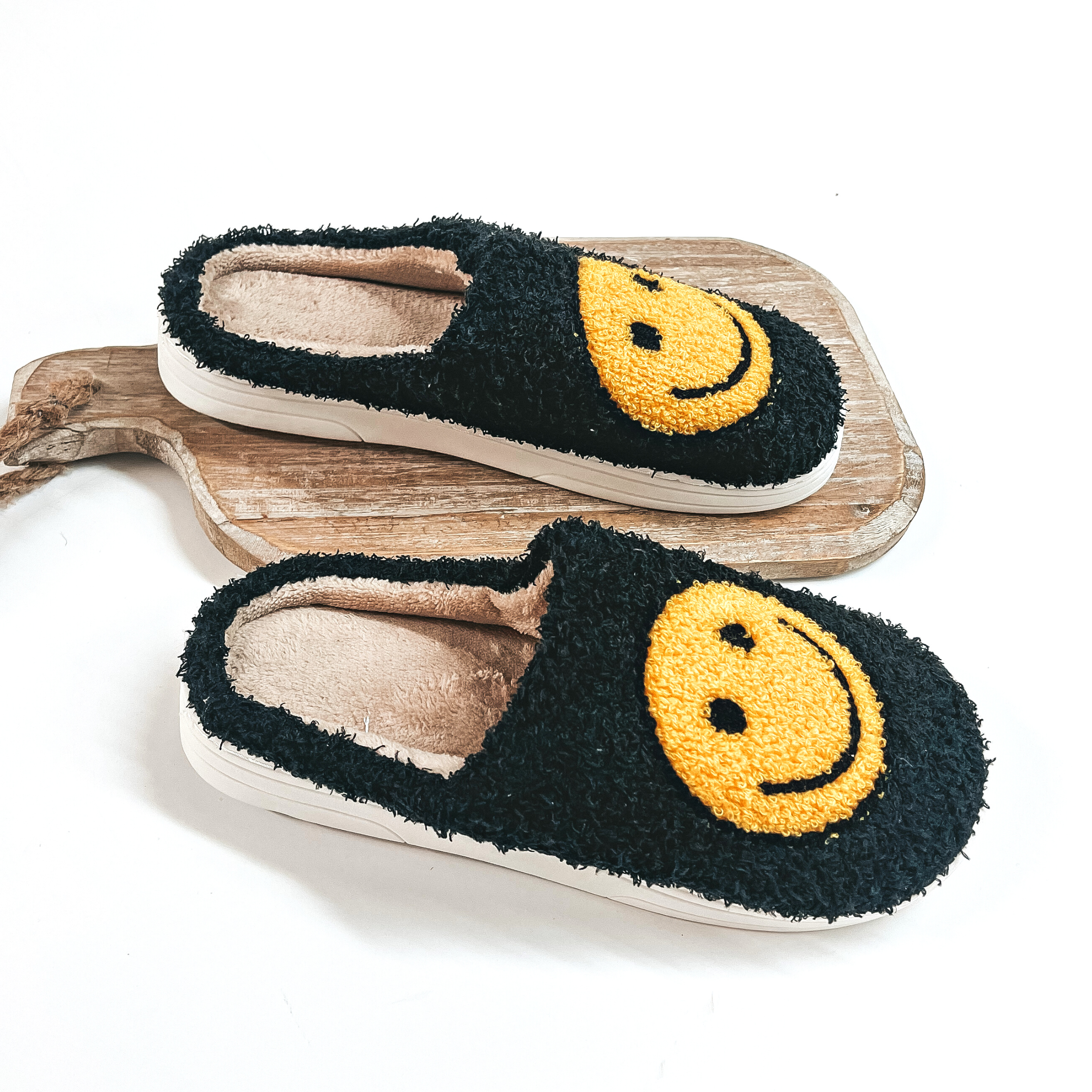 There are sherpa slippers in black with yellow happy faces on top of each  slipper. The inside is in brown and the bottom sole is white. One slipper  is placed on a wooden slab and the other slipper is on a white background.